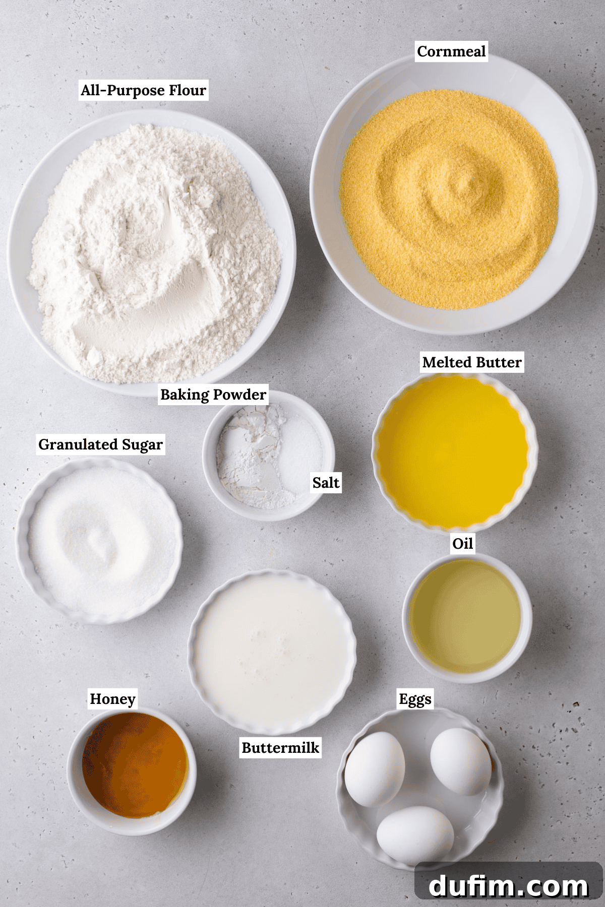 A collection of corn muffin ingredients in bowls on a countertop: all-purpose flour, cornmeal, granulated sugar, salt, baking powder, melted butter, oil, honey, buttermilk, and three eggs. Each item is labeled with text.