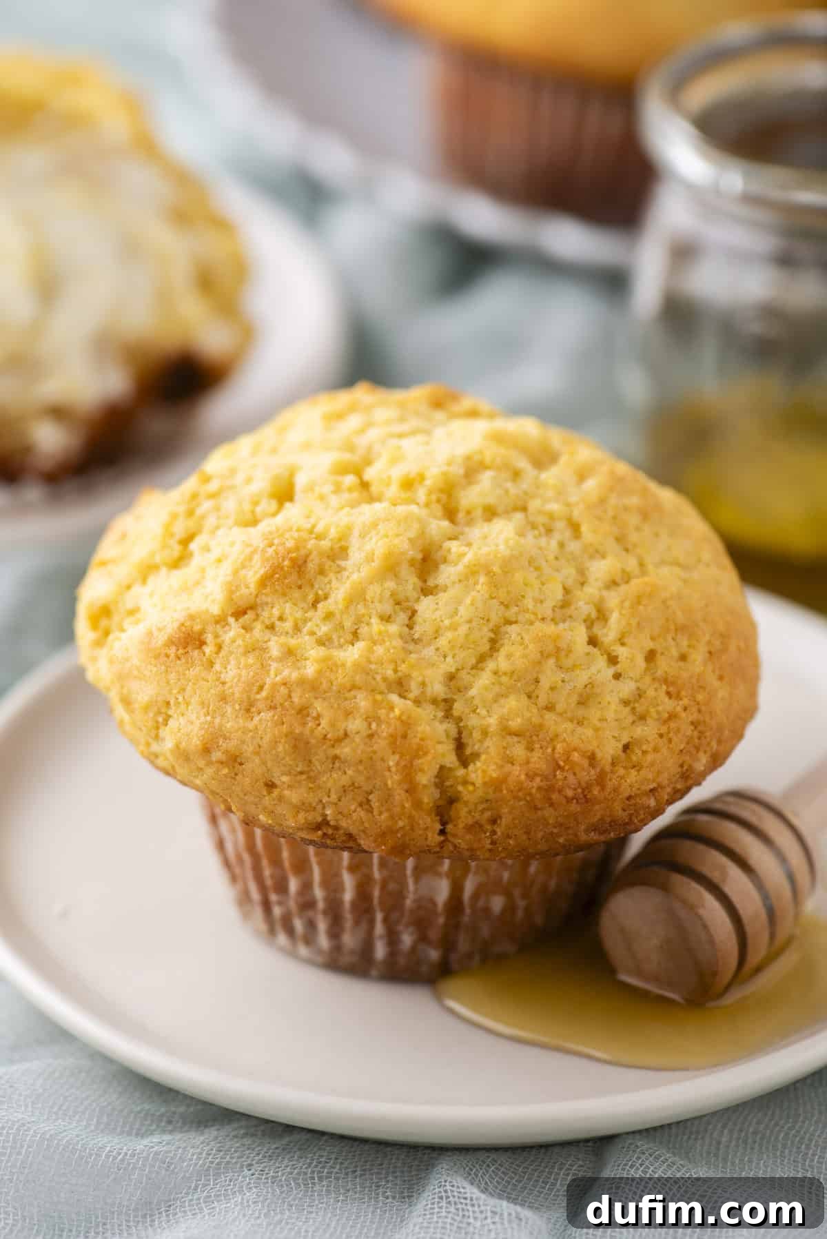 A golden-brown muffin on a white plate with a wooden honey dipper coated in honey beside it. The background shows a glass jar of honey and another plate with a partially visible muffin. The setting conveys a cozy, homemade feel.