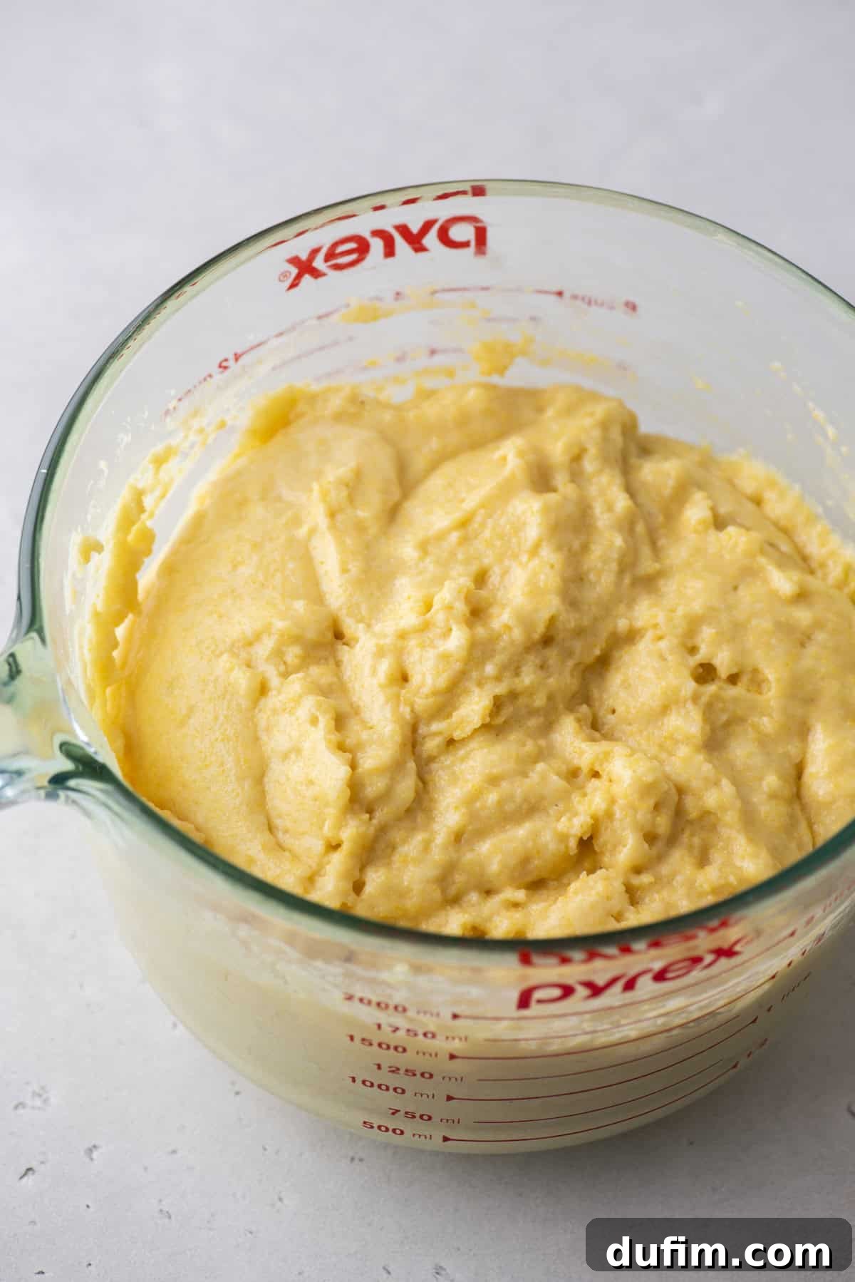 A clear glass measuring cup filled with light yellow, thick cornbread batter. The sides of the cup display red measurement markings. The background is a light-colored surface.