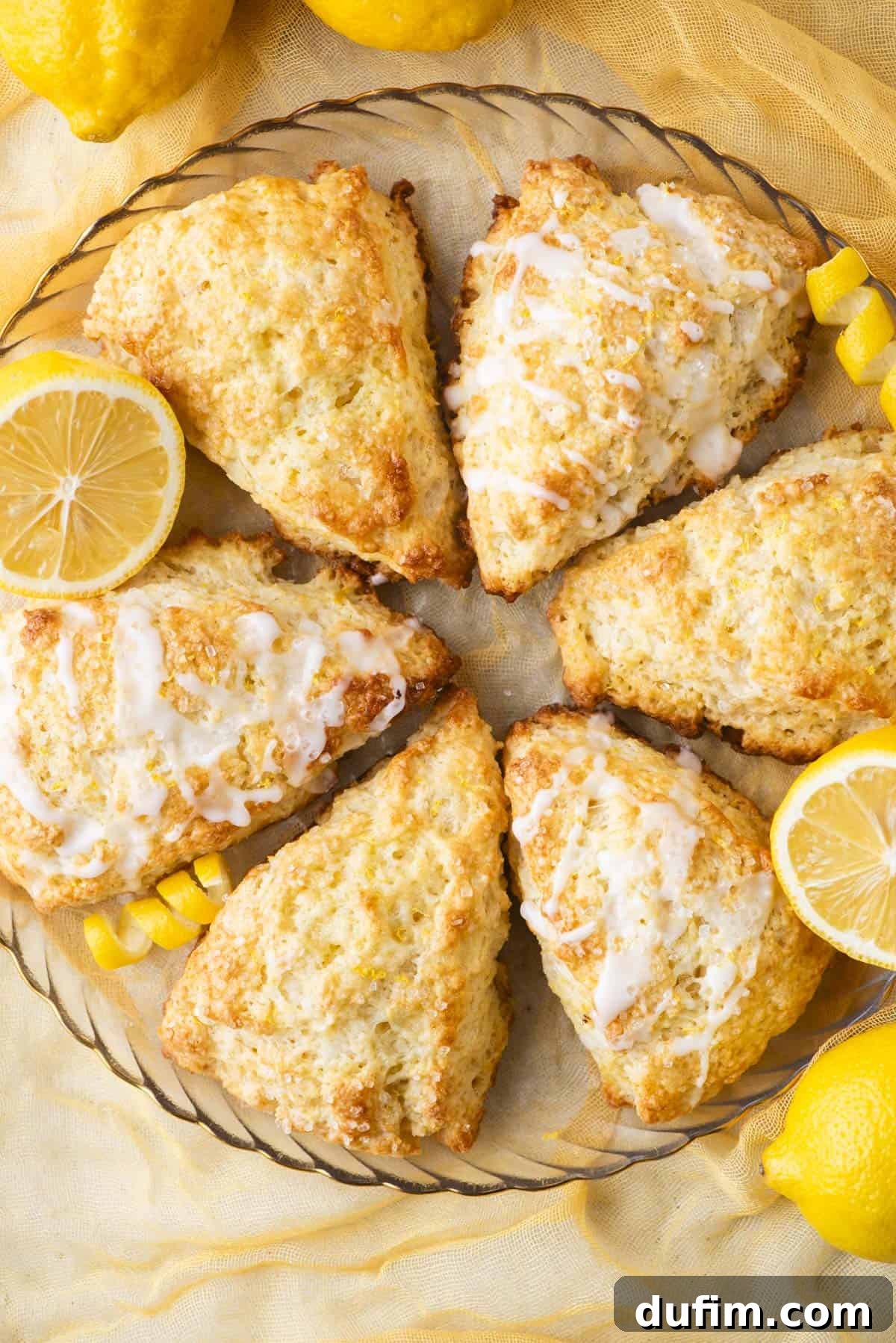 A plate of six golden-brown lemon scones arranged in a circle, drizzled with white icing and garnished with fresh lemon slices. Bright yellow lemons surround the plate on a light yellow cloth background, creating a vibrant and inviting scene.