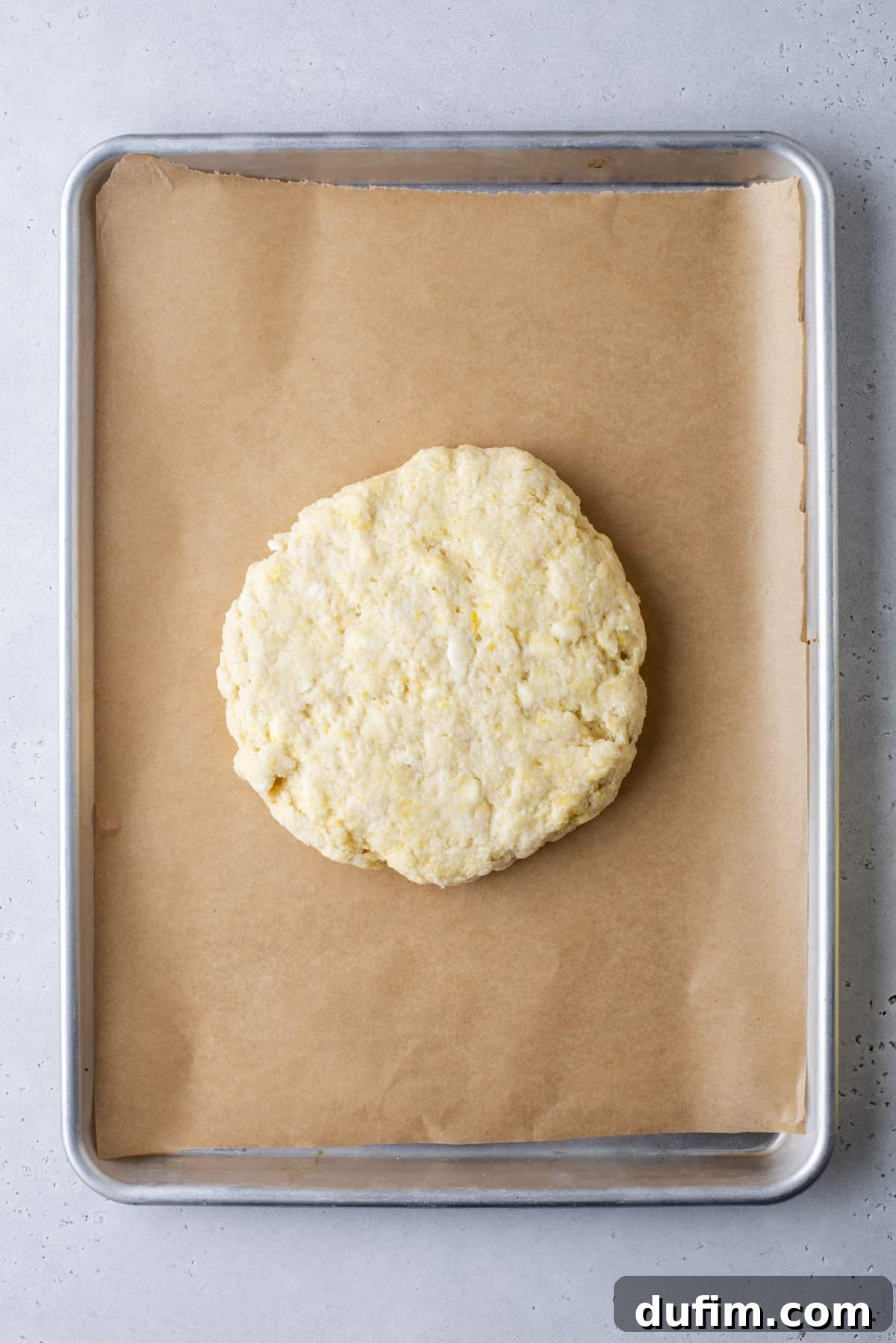A round, uncooked scone dough rests on a sheet of parchment paper, carefully placed on a metal baking tray. The dough's surface has a subtle texture, and the tray sits on a light gray countertop, ready for the next stage of preparation.
