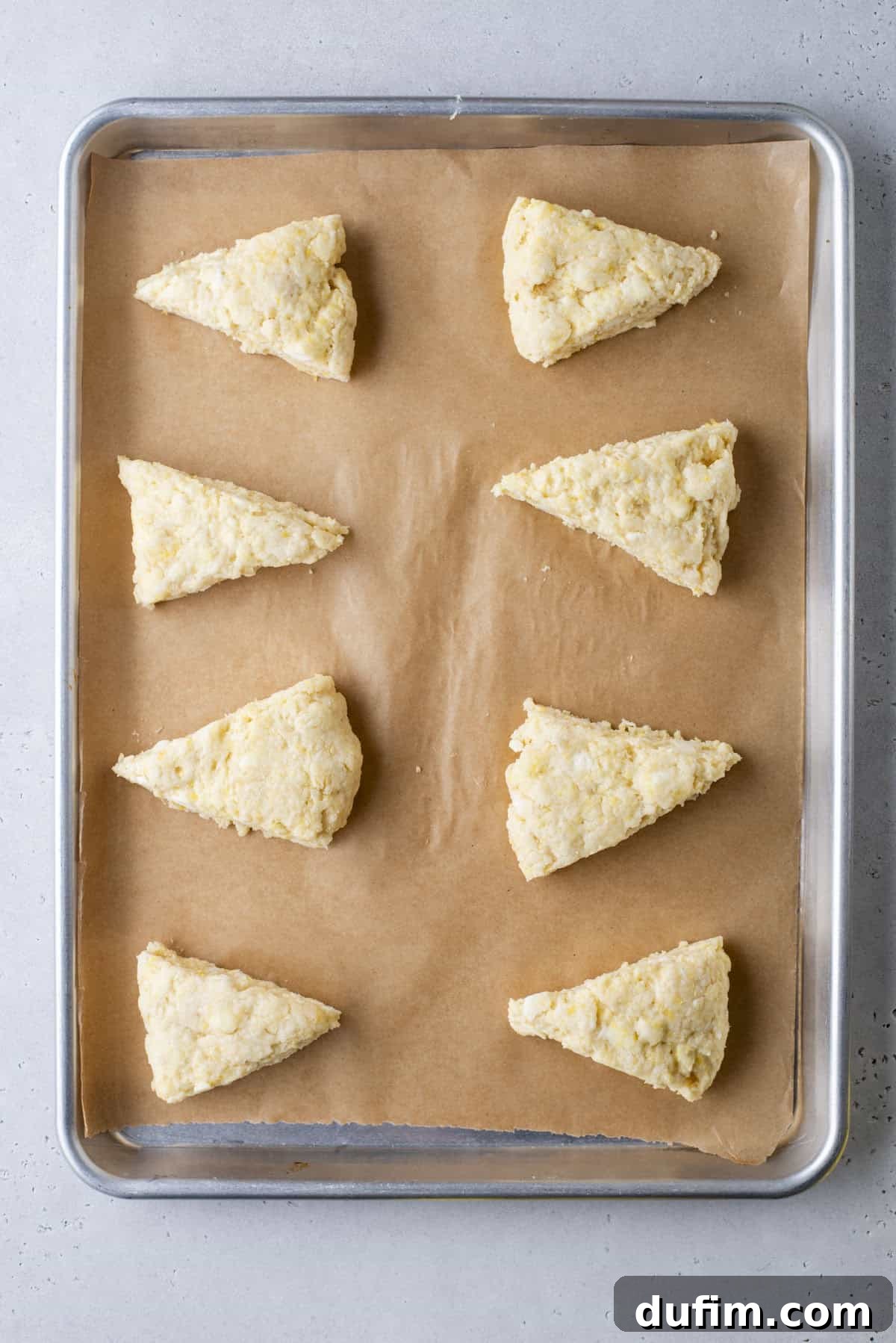 A baking sheet neatly holds eight unbaked, triangular scones, each spaced evenly on a piece of parchment paper. The dough appears pale and subtly textured, awaiting its transformation in the oven, positioned on a light gray countertop.