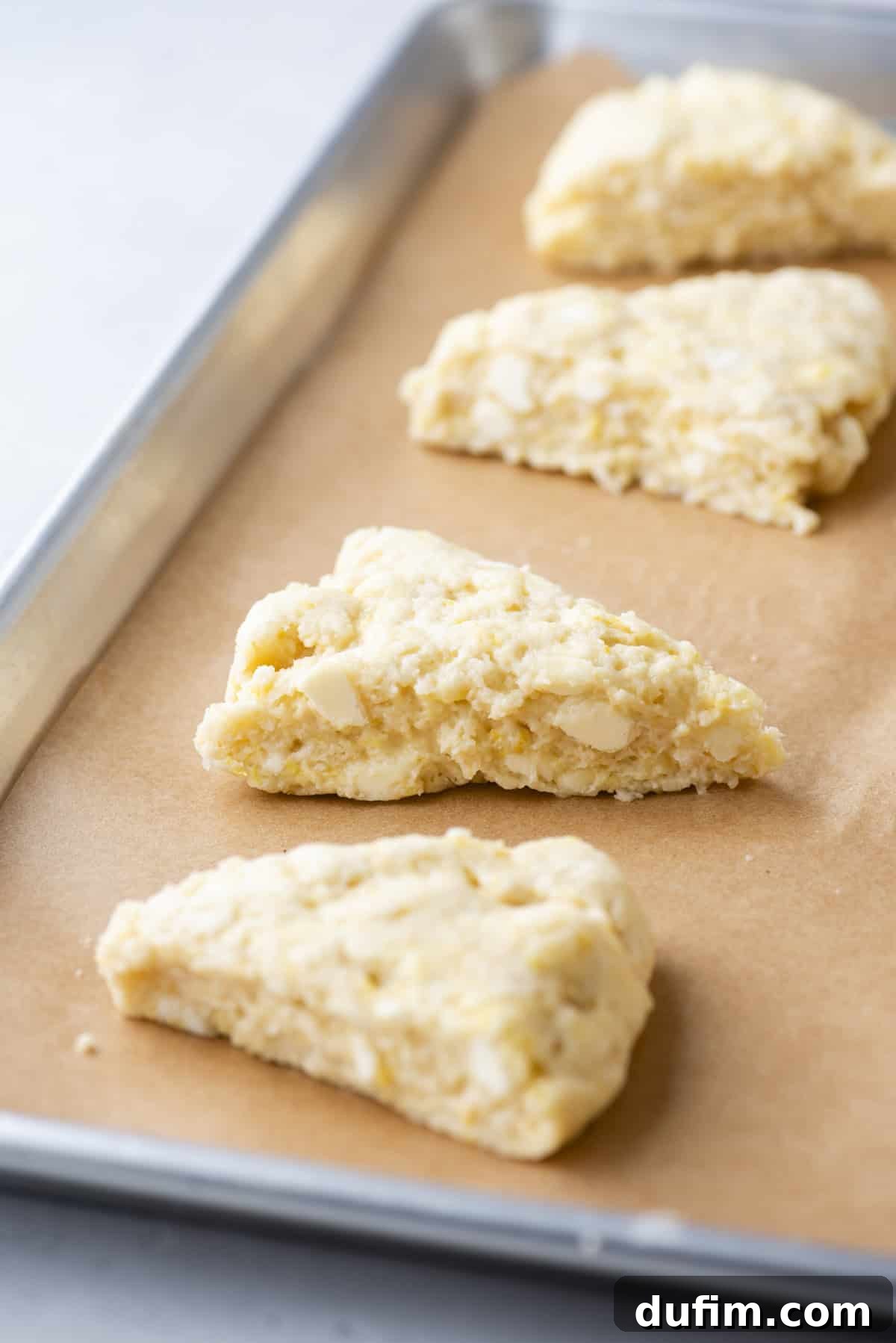 Four unbaked scones are arranged on a parchment-lined baking sheet, spaced appropriately. The scones display a light, somewhat crumbly texture, with distinct chunks of unbaked dough visible, indicative of the cold butter technique.