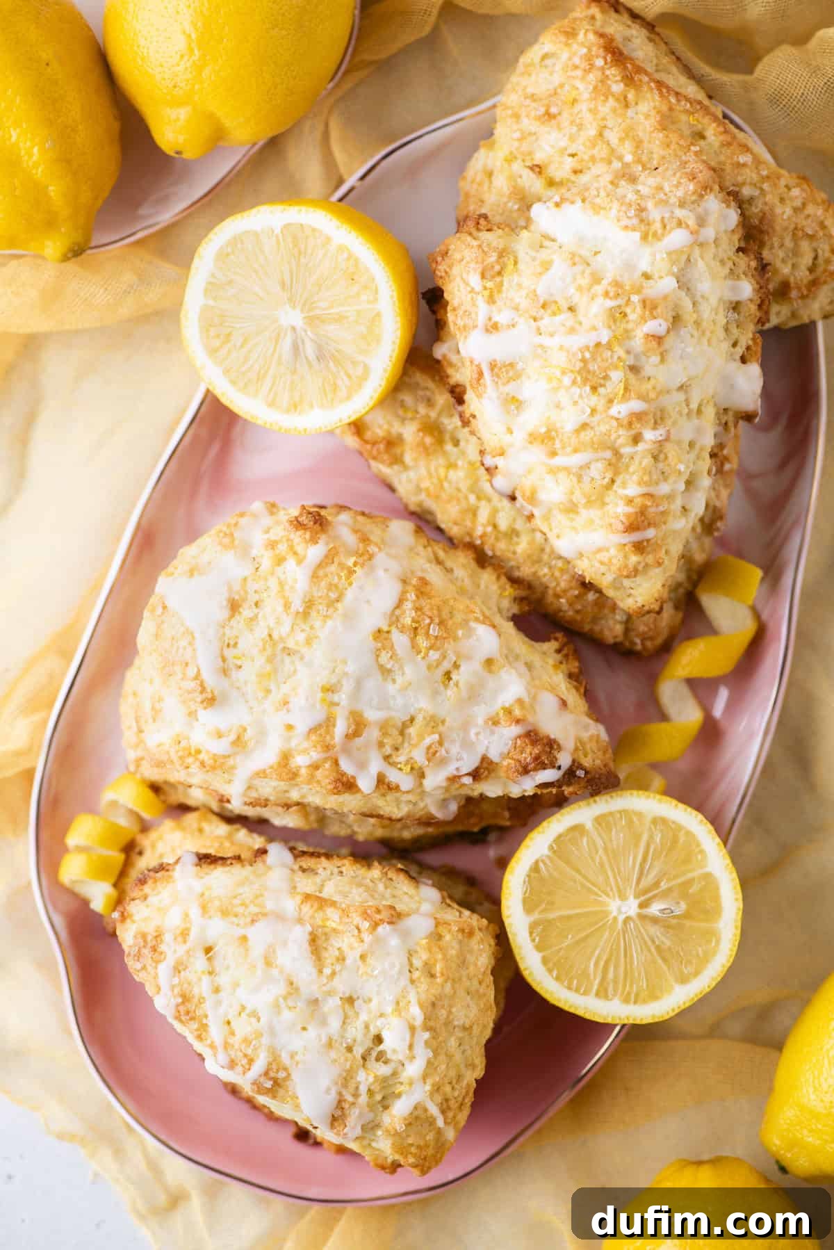 A vibrant plate of lemon scones generously drizzled with white icing, beautifully surrounded by whole and halved fresh lemons. The scones are artfully arranged on a pink plate atop a sunny yellow cloth, highlighting their golden-brown, textured appearance and the delicate lemon zest garnish, making for an appetizing display.
