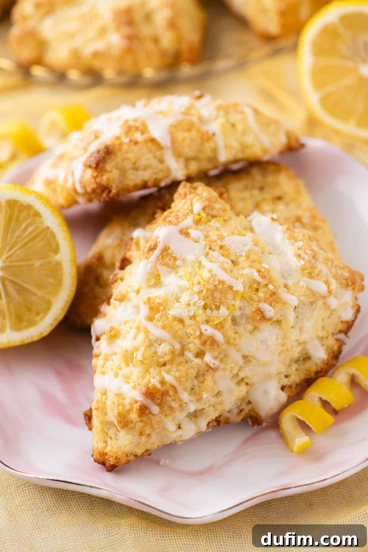 A tastefully arranged plate showcasing three triangular lemon scones, each elegantly drizzled with white icing and artfully garnished with fresh lemon slices and delicate zest curls. The scones boast a rich golden hue and a visibly flaky texture, set against a gentle soft yellow background, enhancing their inviting appeal.