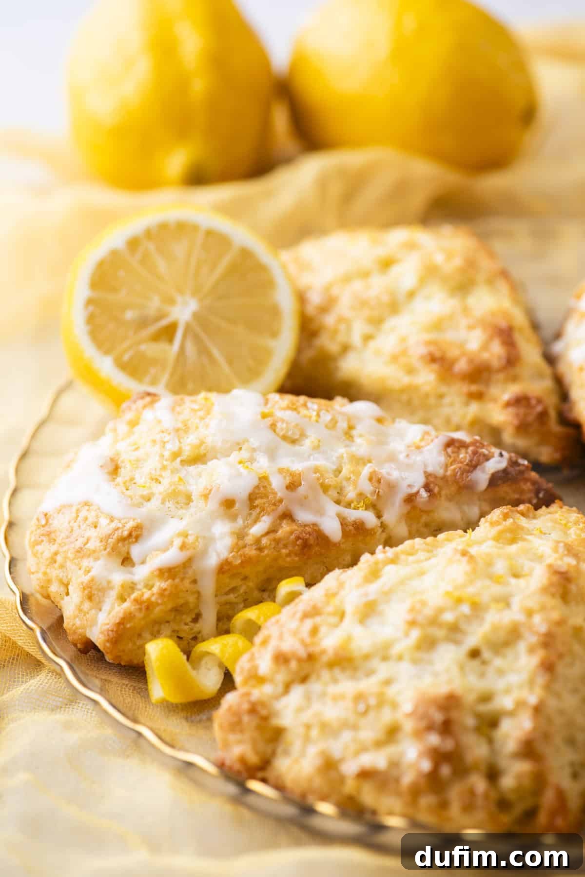 A close-up of beautifully glazed lemon scones resting on a textured plate, garnished with a vibrant lemon slice and delicate lemon zest curls. In the softly blurred background, two whole lemons are placed on a yellow cloth, creating a warm, well-lit scene that emphasizes the scones' inviting texture and fresh citrus appeal.
