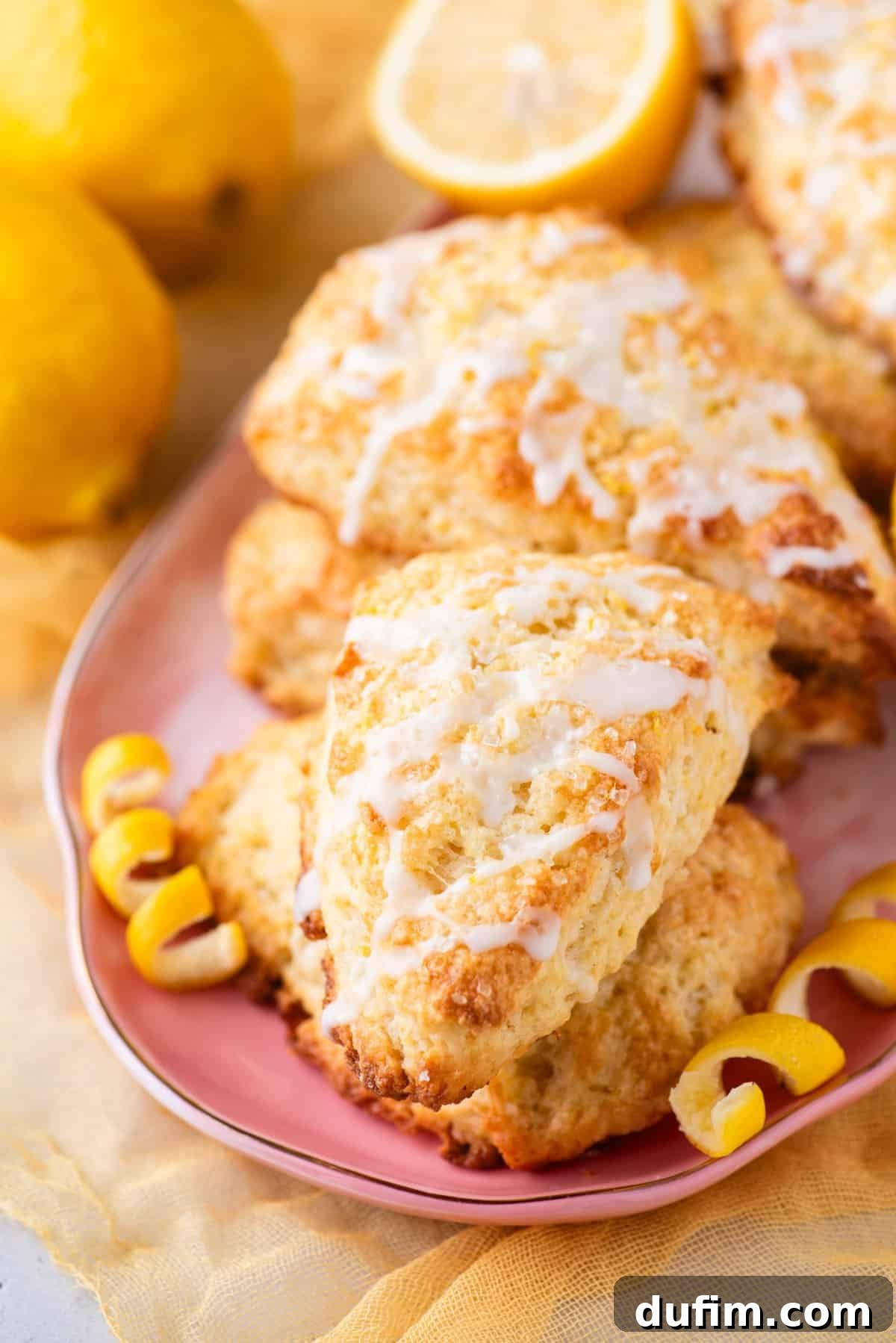 A stack of freshly baked lemon scones, generously drizzled with white icing, gracefully rests on a pink plate. In the background, two whole, vibrant lemons and delicate lemon peel garnishes are visible on a soft yellow cloth, completing a charming and appetizing tableau.