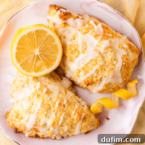 Two lemon scones drizzled with white icing are presented on a plate, accompanied by a halved lemon and a twisted lemon peel garnish. The plate rests on a yellow cloth, creating a bright and inviting image.