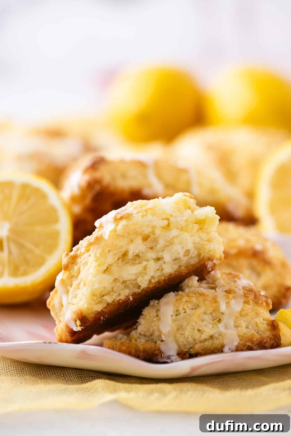 A close-up shot of glazed lemon scones, stacked invitingly on a white plate. The scones exhibit a delightful, crumbly texture with visible traces of the sweet, white icing. In the softly blurred background, vibrant yellow lemons enhance the fresh, sunny ambiance of the scene.