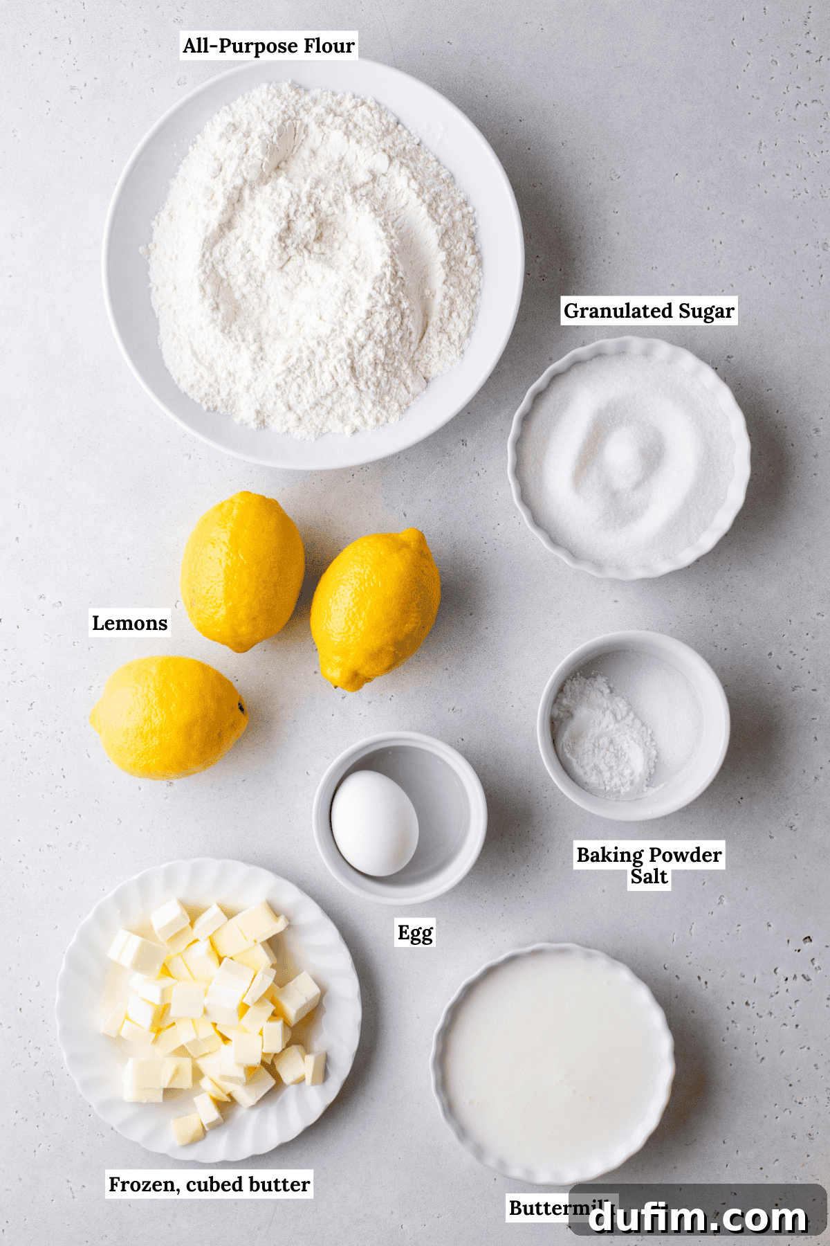 Essential ingredients for lemon scones are neatly laid out on a table, including all-purpose flour, granulated sugar, fresh lemons, a single egg, frozen cubed unsalted butter, and buttermilk. Small dishes hold baking powder and salt, completing the display for an organized baking session.