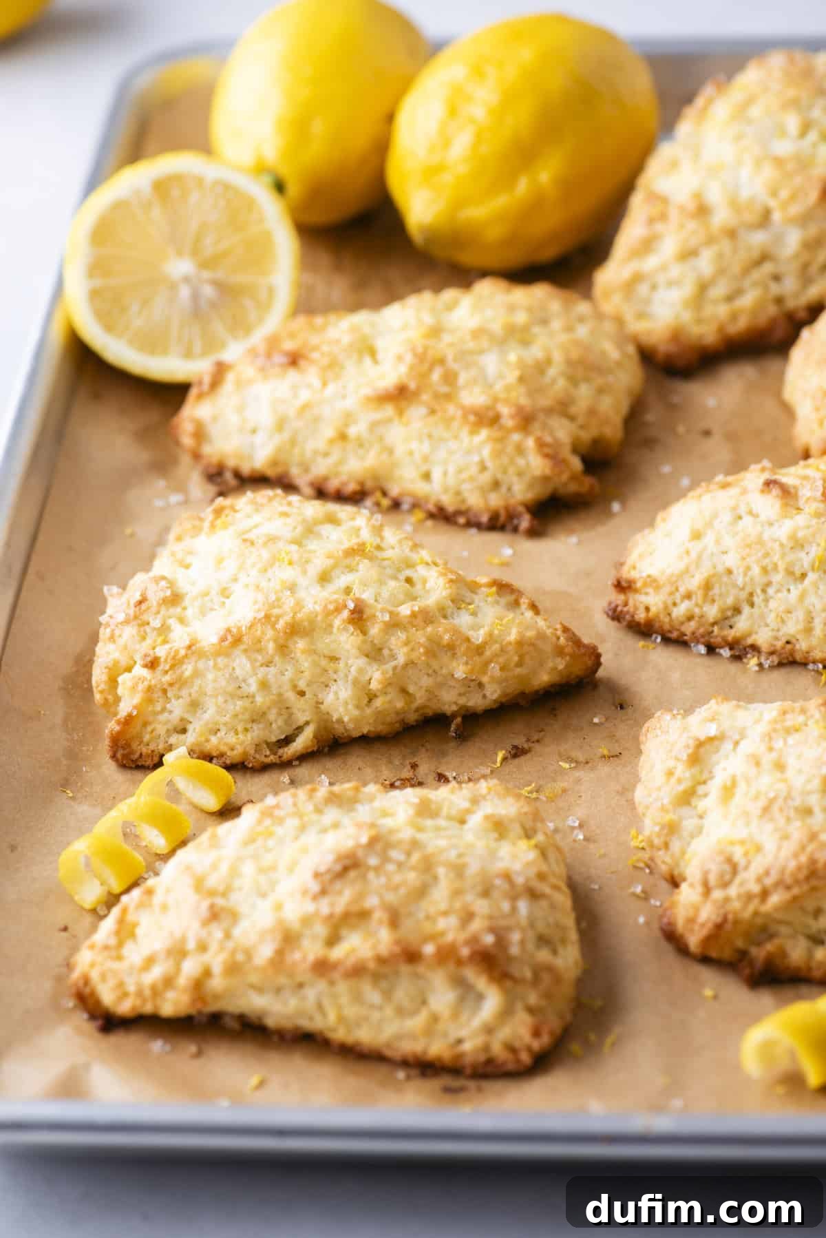 A parchment-lined baking tray holds an array of golden, triangular lemon scones, perfectly spaced and ready for baking. Fresh lemons, a vibrant lemon slice, and delicate curls of lemon zest are artfully arranged in the background, adding to the fresh and appealing presentation.