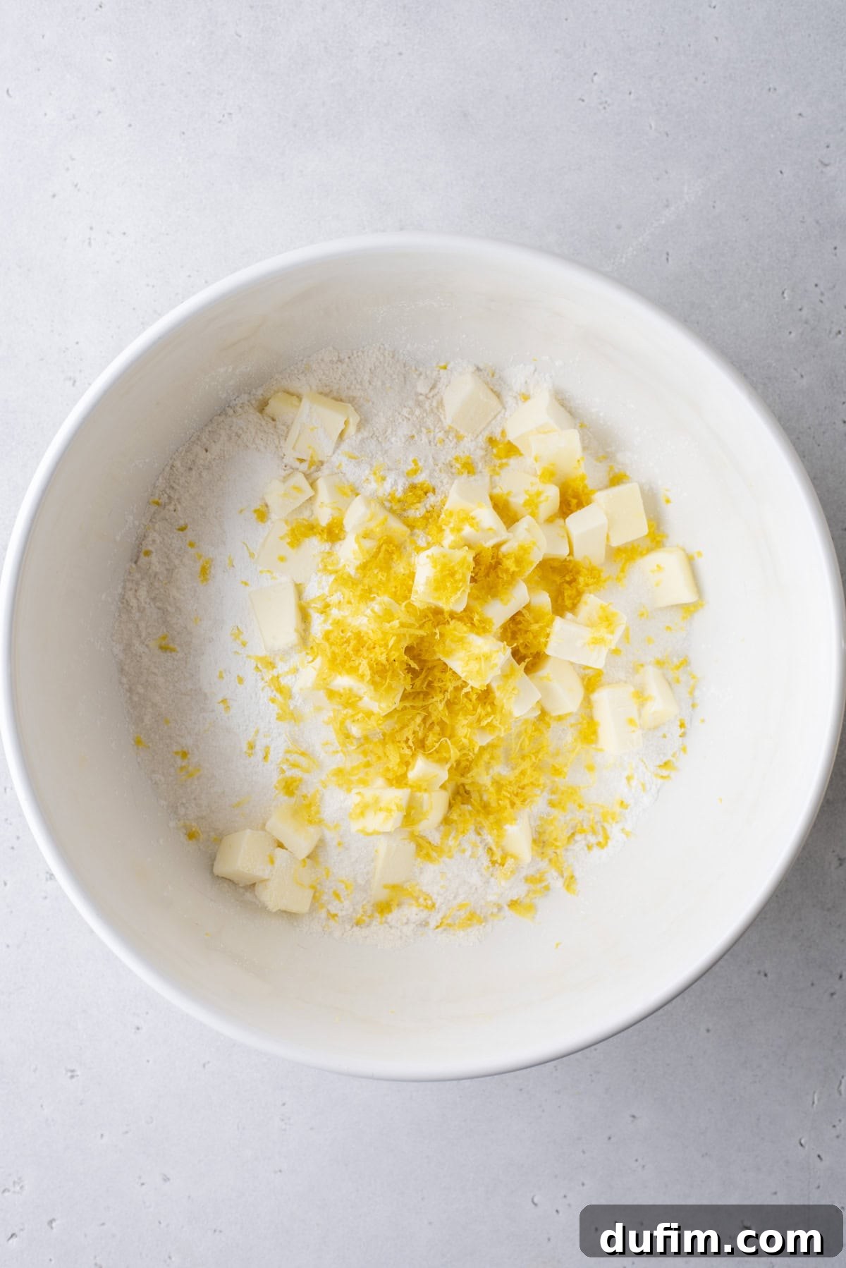 A white bowl on a light gray surface, containing precise cubes of cold butter, finely grated lemon zest, and all-purpose flour. The ingredients are perfectly prepped for the initial mixing stage of a baking recipe, highlighting attention to detail.