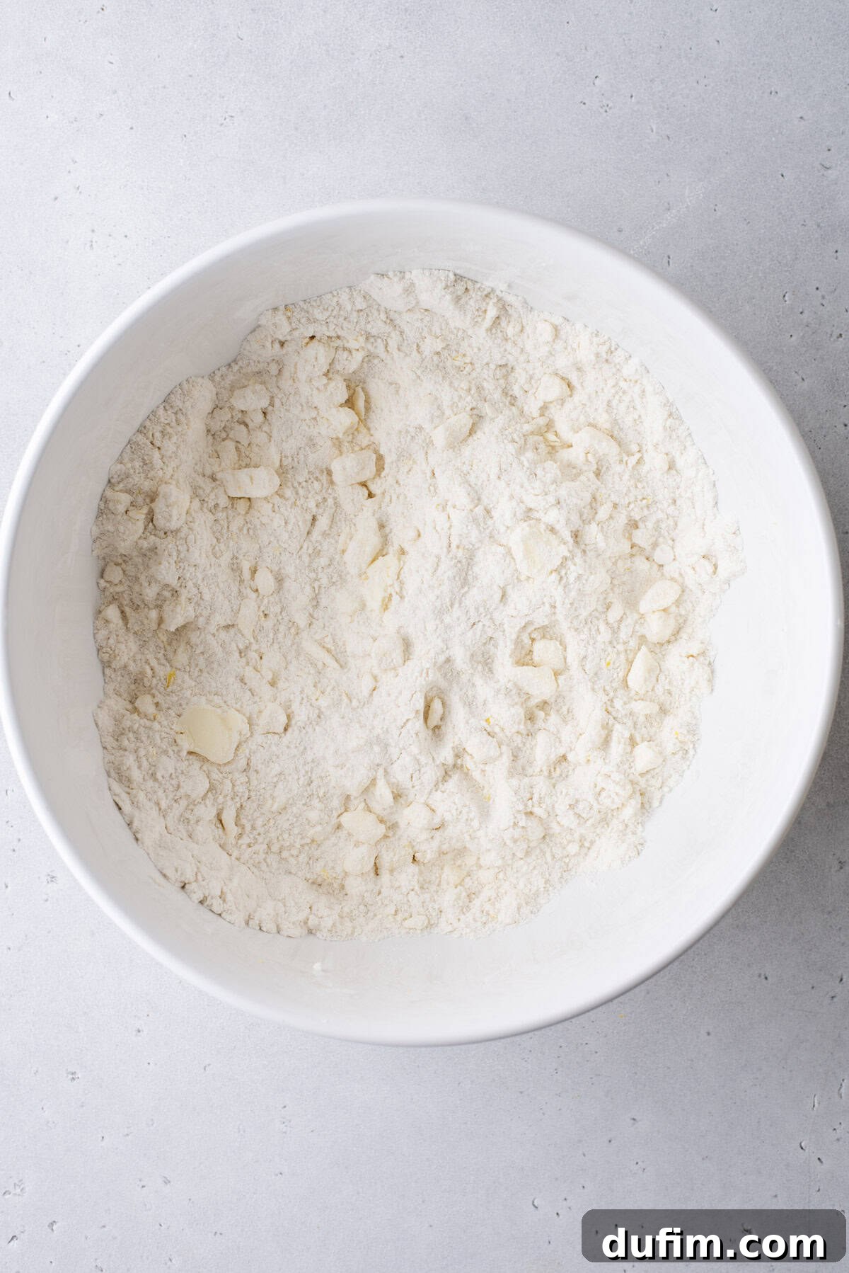 A white mixing bowl on a light gray surface, holding a partially combined mixture of flour and small, distinct pieces of butter. This image captures the crucial initial stages of creating a scone or pastry dough, showcasing the proper 'cut in' technique.