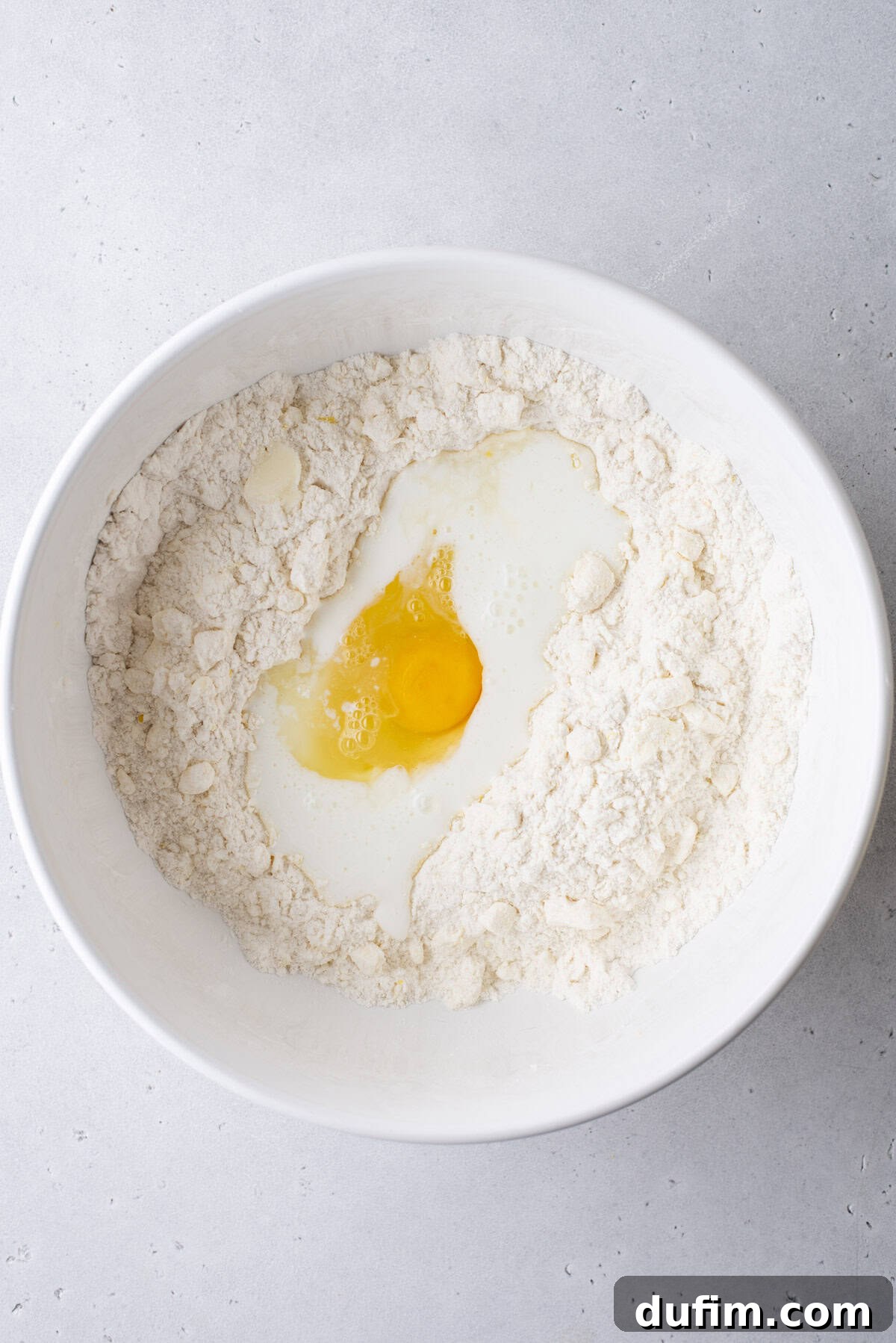 A white bowl with a central well containing flour, buttermilk, and a cracked egg, poised for mixing. The bowl rests on a clean, light gray surface, indicating the preparation stage for combining wet ingredients into the dry mixture.