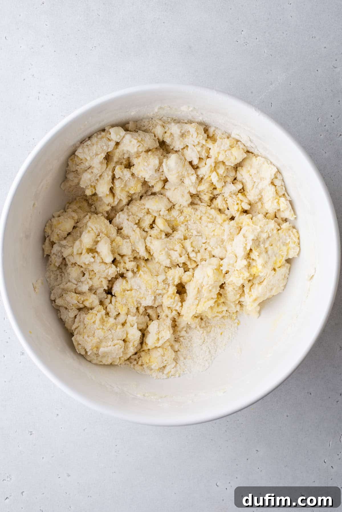 A white bowl contains a light-colored, crumbly dough, likely a scone or pastry dough, resting on a smooth, light gray surface. The texture suggests minimal mixing, preserving the cold butter pieces for flakiness.