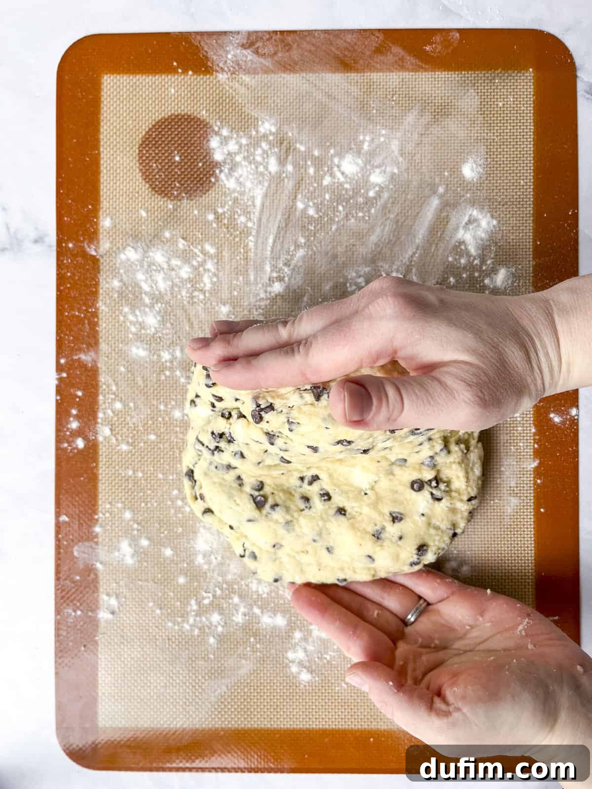Hands are gently shaping chocolate chip cookie dough on a floured silicone baking mat. The focus is on the careful handling of the dough to prepare it for baking, illustrating a key step in the recipe.