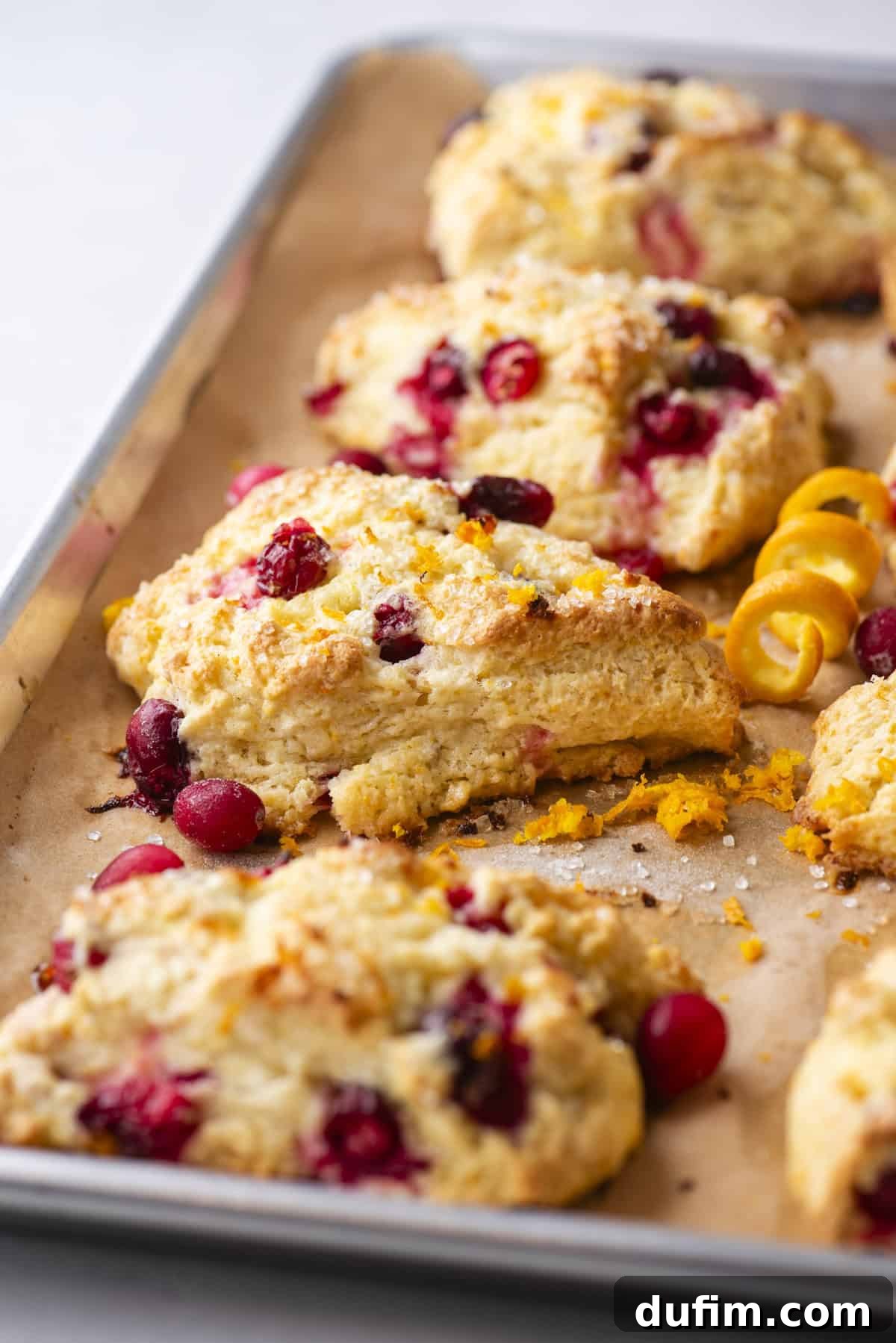 Zesty Cranberry Orange Scones 2 Close-up of freshly baked cranberry orange scones on a parchment-lined baking sheet. The scones are golden brown with visible cranberries and sprinkled sugar on top, highlighting their flaky texture. A few orange zest curls and a whole orange peel add a fresh, vibrant touch to the side.