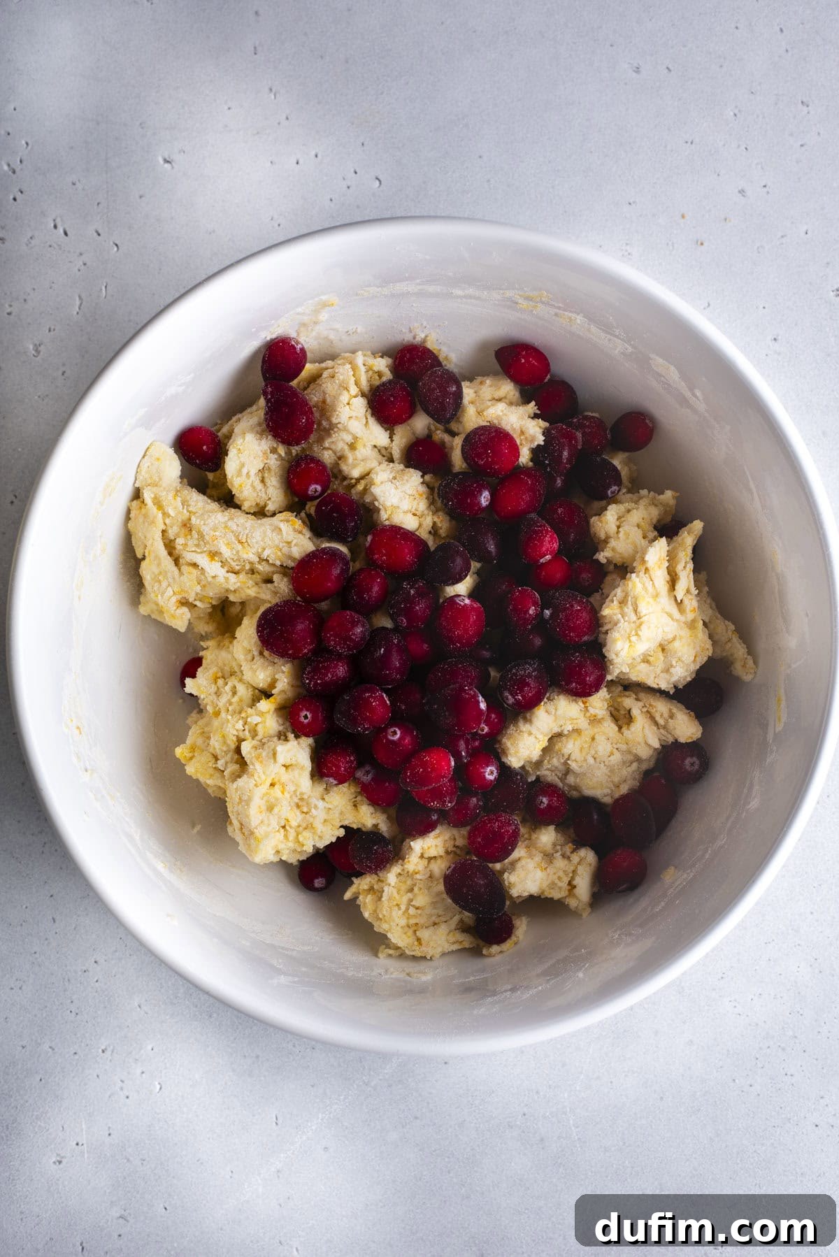 Zesty Cranberry Orange Scones 12 A white bowl holds a partially mixed scone dough with vibrant red fresh cranberries scattered on top. The dough appears soft and a bit crumbly, ready for final incorporation, showcasing the key fruit ingredient.