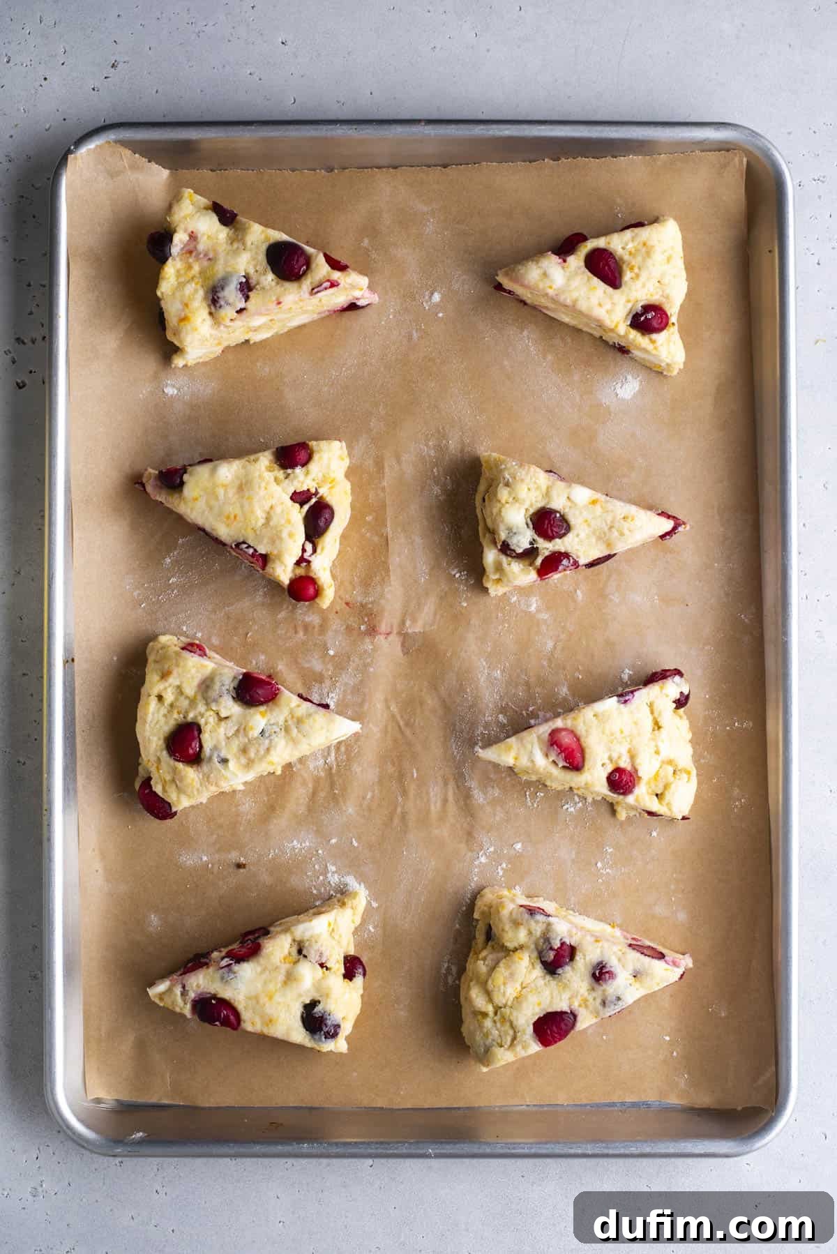 Zesty Cranberry Orange Scones 14 Eight unbaked, triangular scone wedges, visibly speckled with cranberries, are neatly arranged and evenly spaced on a parchment-lined baking sheet. The rustic, homemade appearance of the dough is ready for chilling and baking. The baking sheet is placed on a light gray surface.