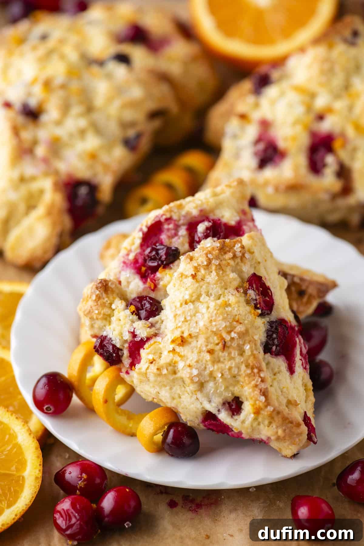 Zesty Cranberry Orange Scones 18 A charming white plate holds several golden-brown cranberry orange scones, delicately topped with sparkling sugar crystals. Fresh, plump cranberries and aromatic orange peel curls are artfully scattered around the plate, enhancing the vibrant color and inviting aroma. More scones and bright orange slices are blurred in the background, creating a warm and appealing scene, perfect for breakfast or dessert.