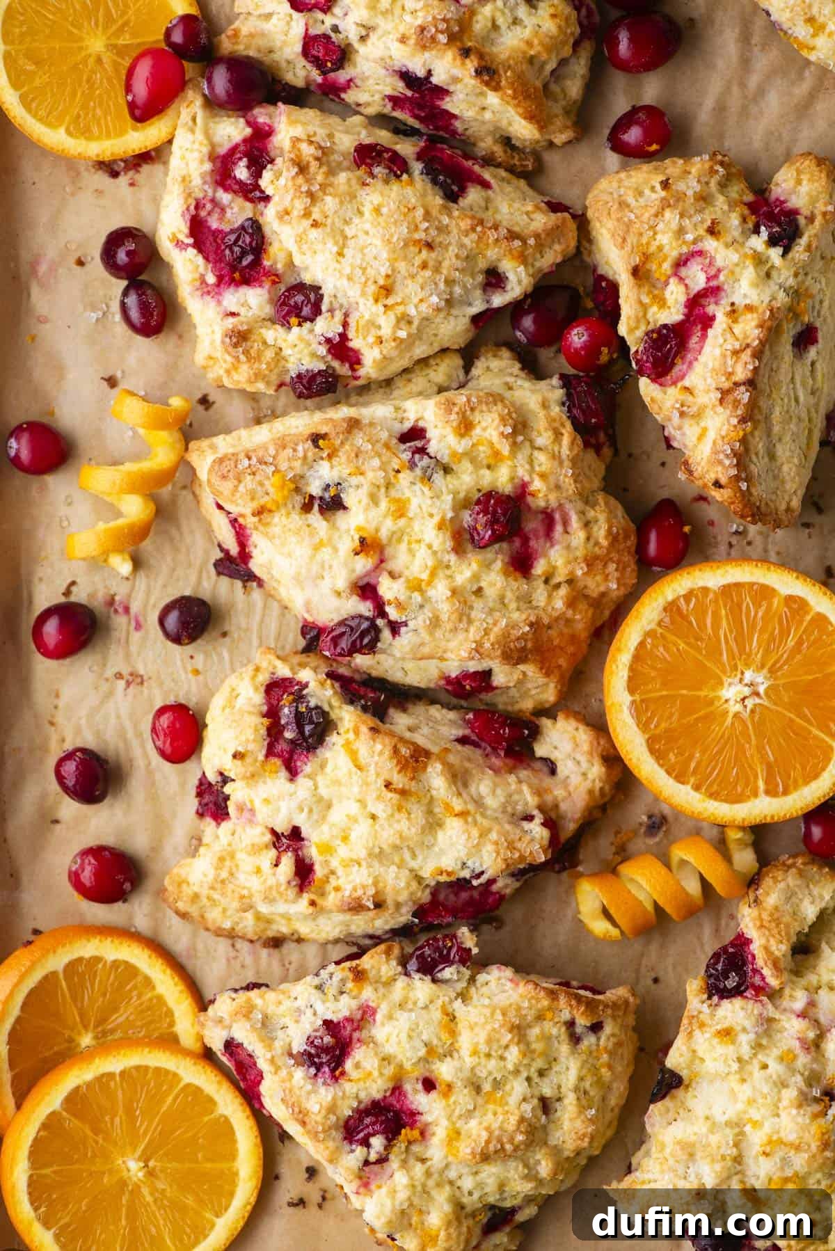 Zesty Cranberry Orange Scones 3 Triangular cranberry-orange scones artfully arranged on a parchment-lined surface, surrounded by vibrant fresh cranberries and bright orange slices. The golden-brown scones showcase visible pieces of tart cranberry and fragrant orange zest, creating an inviting and colorful display of freshly baked goods.