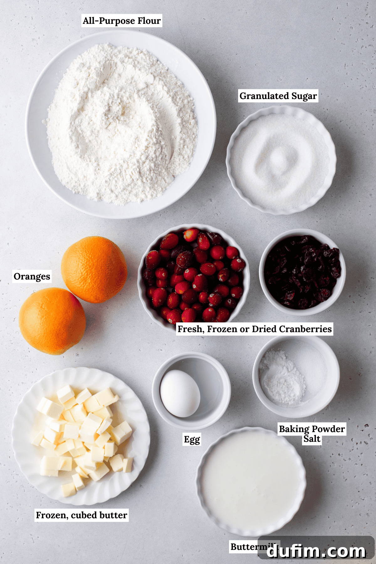 Zesty Cranberry Orange Scones 4 An inviting overhead view of all the fresh ingredients meticulously laid out for baking cranberry orange scones. The display includes bowls filled with all-purpose flour, glistening granulated sugar, plump cranberries, perfectly cubed cold butter, a farm-fresh egg, creamy buttermilk, whole oranges, and a small dish containing baking powder and salt, all arranged on a bright, clean surface.