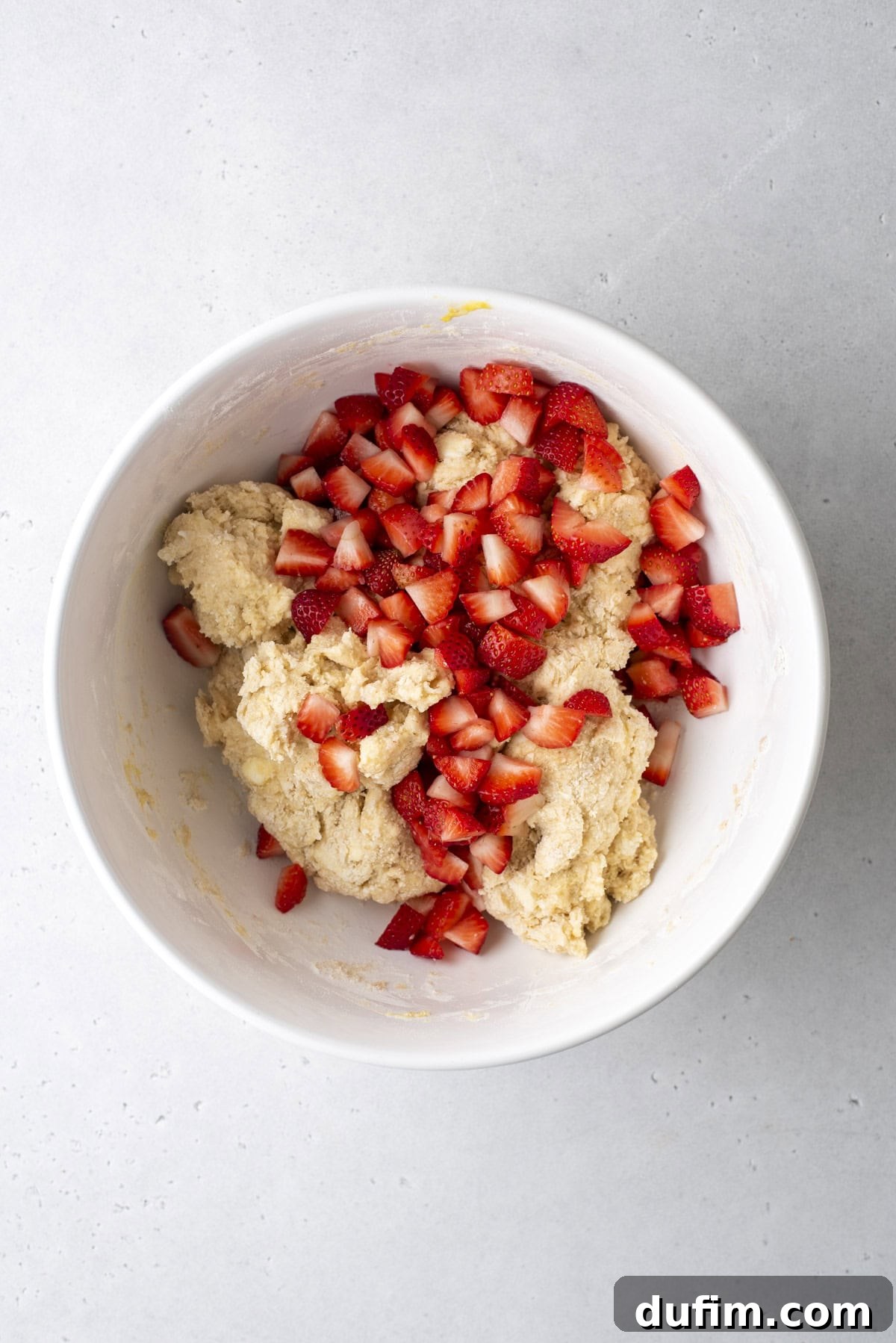 Juicy Strawberry Scones 11 A white bowl contains dough and chopped strawberries on a light gray surface. The dough is a light color, while the diced strawberries, bright red with hints of white, are scattered on top of the mixture.