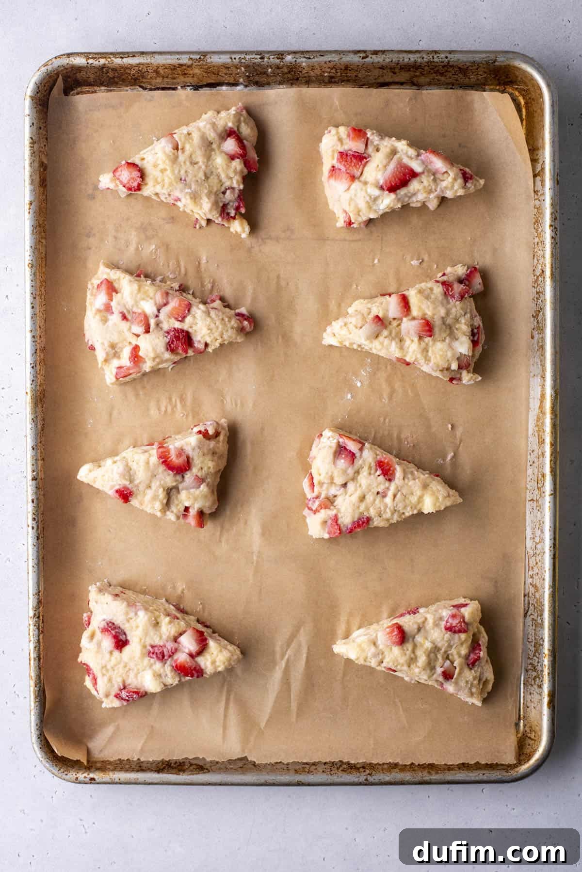 Juicy Strawberry Scones 15 Eight unbaked strawberry scones are arranged on a parchment-lined baking sheet. The scones are triangular with visible strawberry chunks, ready for baking.