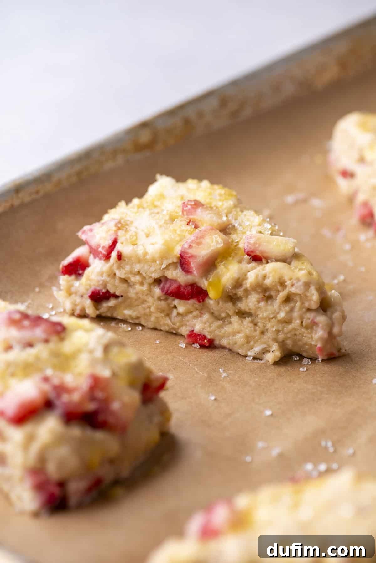 Juicy Strawberry Scones 16 Close-up of a triangular strawberry scone on a parchment-lined baking sheet. The scone is golden-brown with visible chunks of strawberries and a sprinkle of sugar on top. Other scones are partially visible in the background.