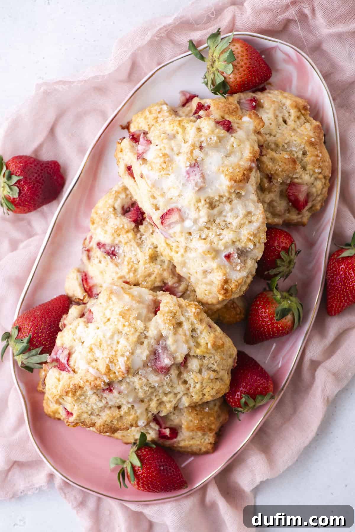 Juicy Strawberry Scones 18 A plate of strawberry scones topped with icing, surrounded by fresh strawberries on a pink cloth. The scones are golden brown with visible strawberry pieces, creating a delicious and inviting presentation.