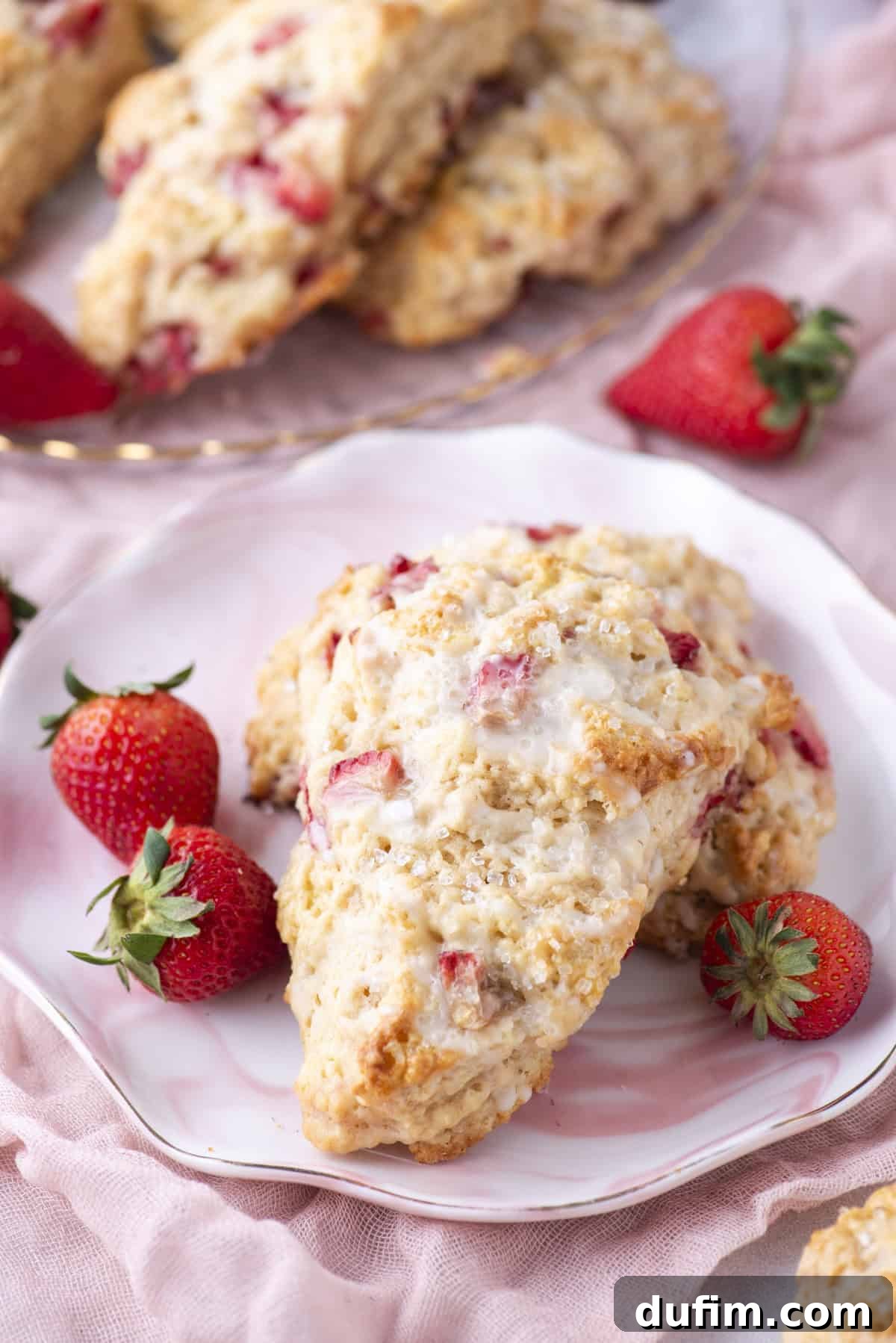 Juicy Strawberry Scones 20 A plate with a strawberry scone garnished with fresh strawberries. The scone has a golden, crumbly texture and visible strawberry pieces. More scones and strawberries are in the background, set on a soft pink fabric.
