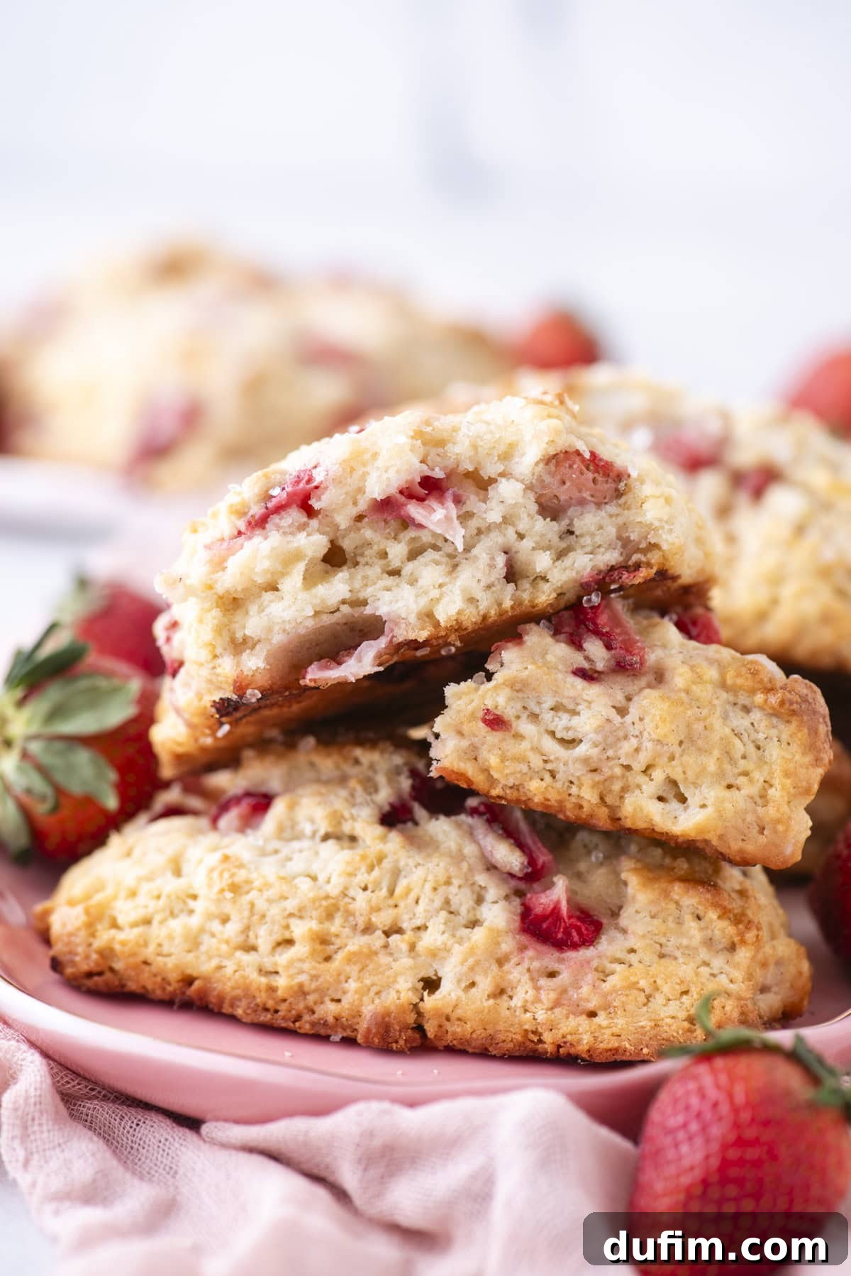 Juicy Strawberry Scones 3 Close-up of freshly baked scones filled with sliced strawberries, stacked on a pink plate. Whole strawberries surround the plate, and a soft pink cloth is partially visible in the foreground.