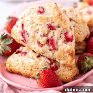 Close-up of two strawberry scones topped with sugar crystals on a pink plate. Fresh strawberries are scattered around the scones, and a light pink cloth is in the background, creating a soft, inviting atmosphere.