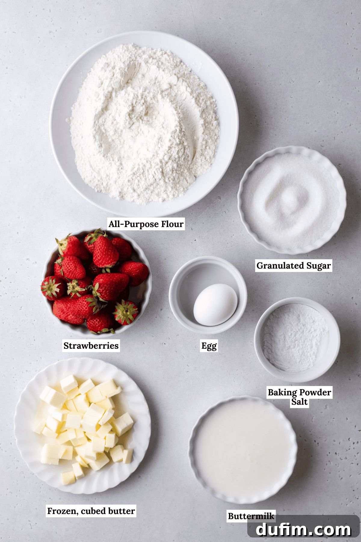 Juicy Strawberry Scones 5 A top view of baking ingredients on a gray surface, including bowls of all-purpose flour, granulated sugar, baking powder, salt, an egg, cubed frozen butter, buttermilk, and fresh strawberries.