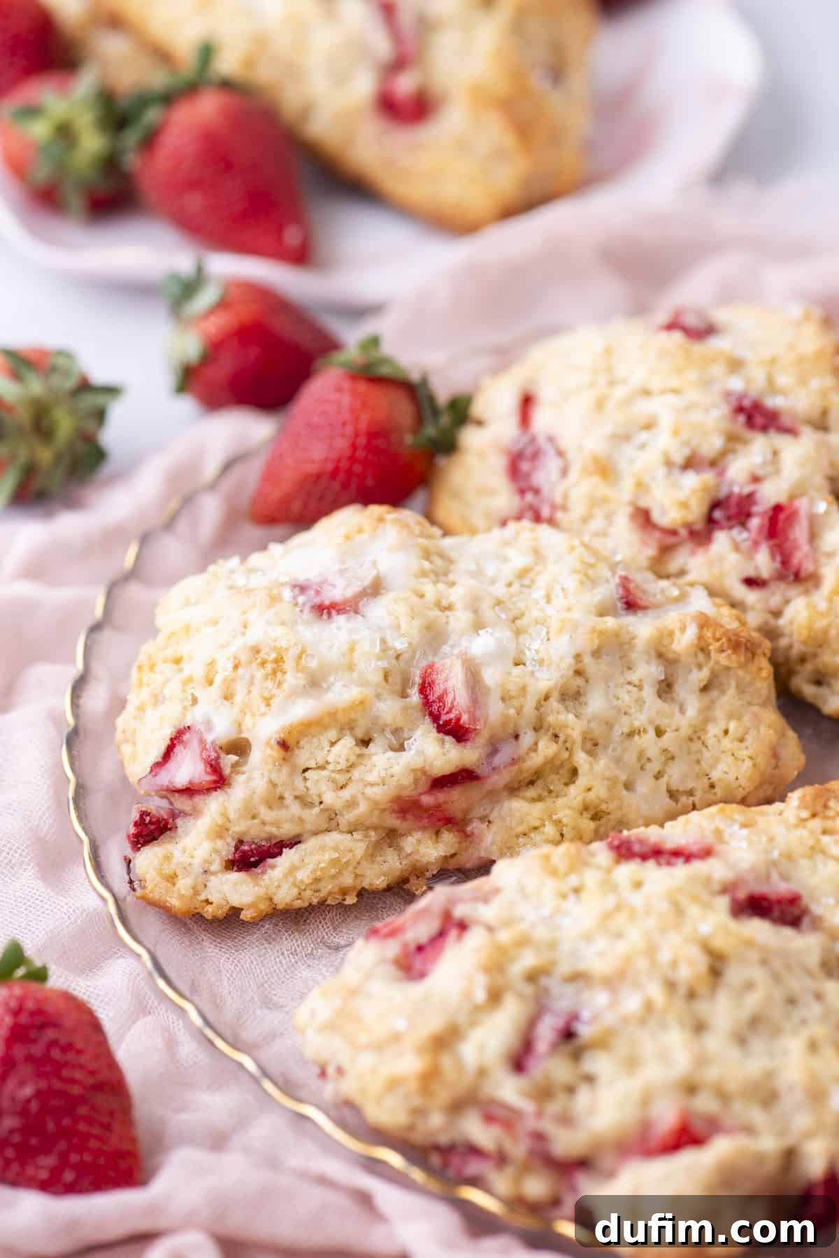 Juicy Strawberry Scones 6 Close-up of freshly baked strawberry scones on a decorative plate. The scones are golden brown with visible strawberry pieces and a light glaze on top. Fresh strawberries are scattered around, and a soft pink cloth is partially visible underneath the plate.