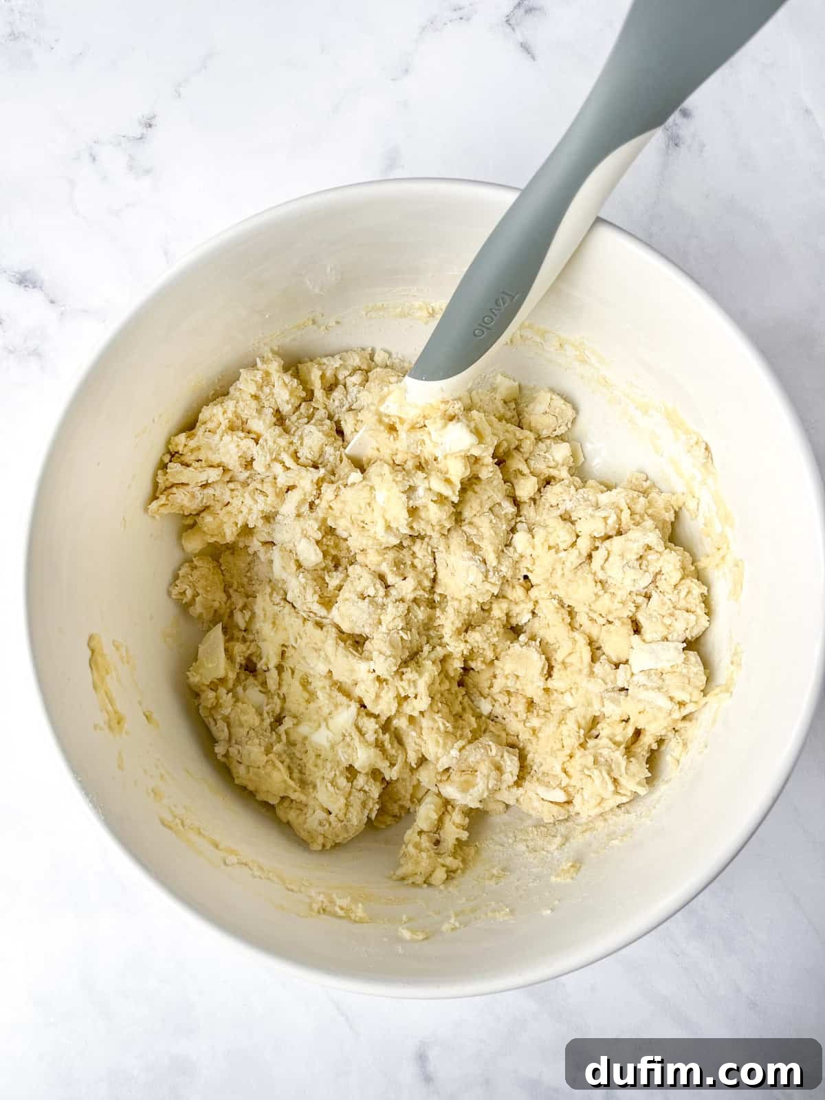 A white mixing bowl filled with crumbly, semi-mixed gluten-free scone dough. A grey spatula rests inside the bowl, which is placed on a marble countertop, showing the dough's sticky consistency.