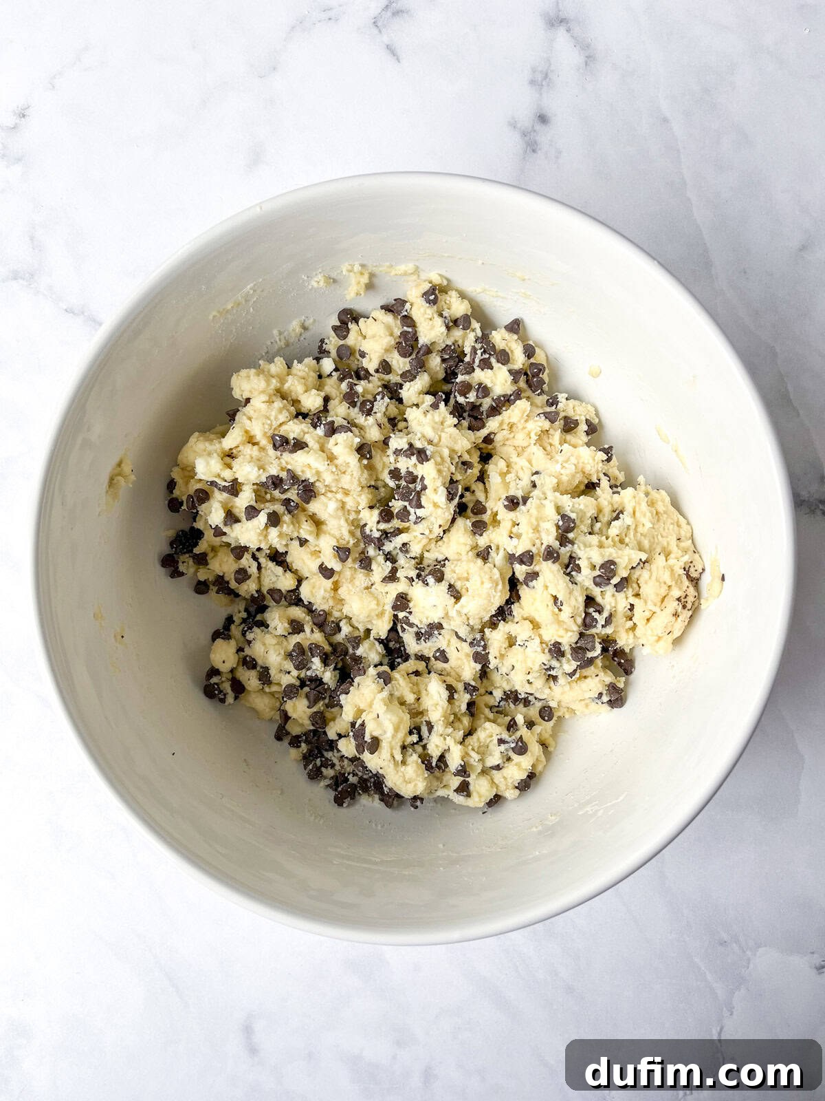 A white bowl filled with chocolate chip scone dough, showing a thick, sticky consistency after wet ingredients and mix-ins have been combined, resting on a light marbled surface.