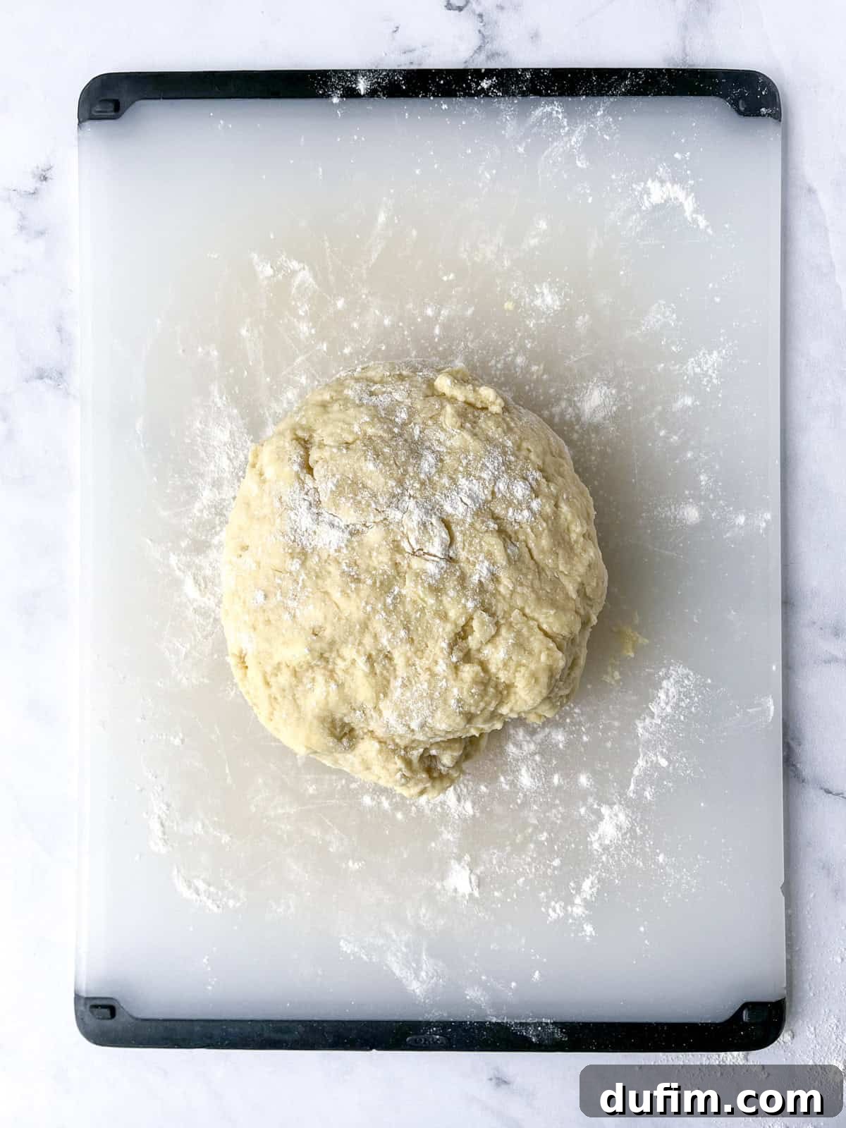 A freshly mixed ball of gluten-free scone dough, lightly dusted with flour, resting on a transparent cutting board with black edges on a marble surface, ready for kneading.