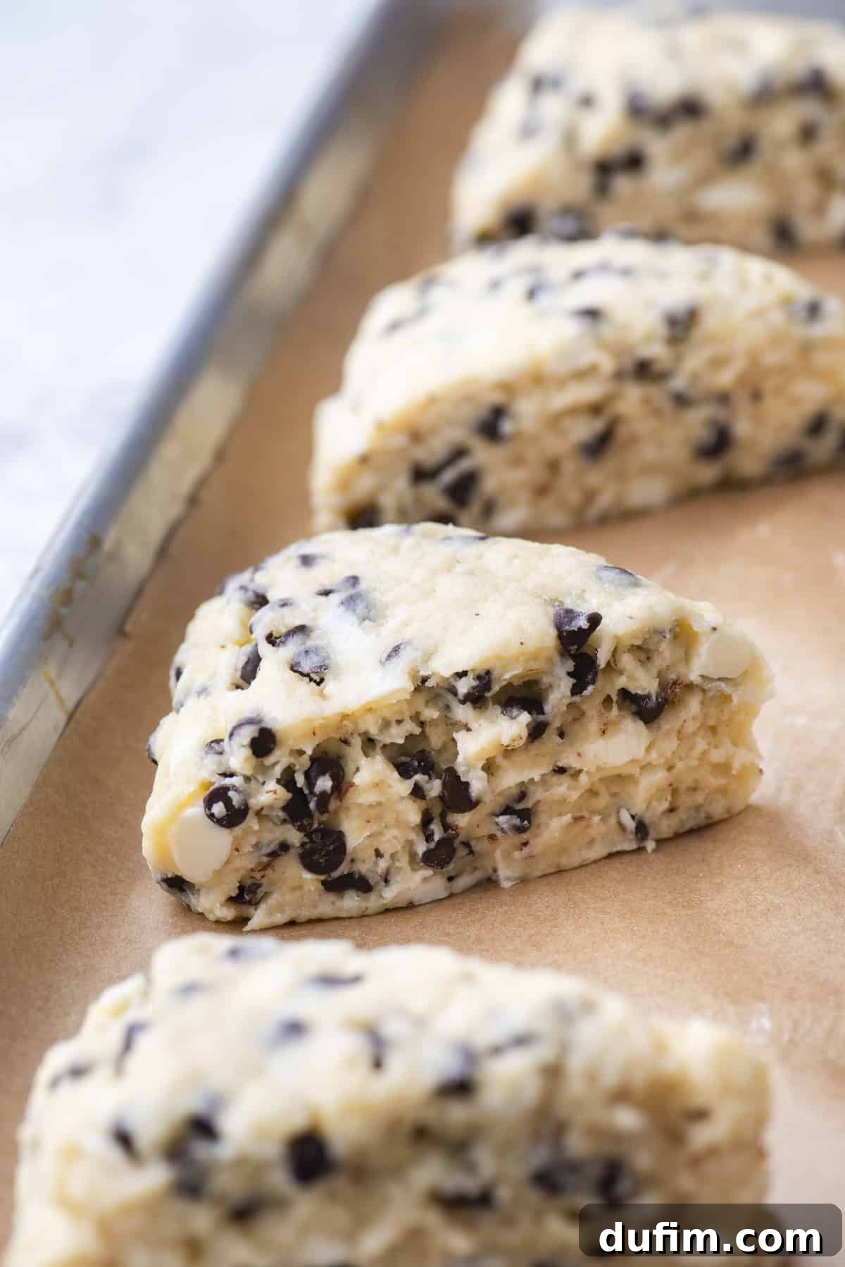 Close-up of four unbaked chocolate chip gluten-free scones on a parchment-lined baking tray. Each scone is triangular, with a light dough speckled with small chocolate chips, showing its readiness for baking.