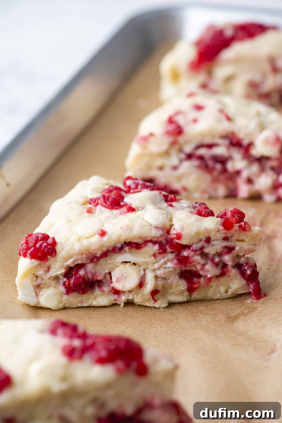 Close-up of freshly baked gluten-free scones featuring raspberries and white chocolate chunks on a parchment-lined baking sheet. The triangular scones display a beautiful flaky texture and vibrant fruit.
