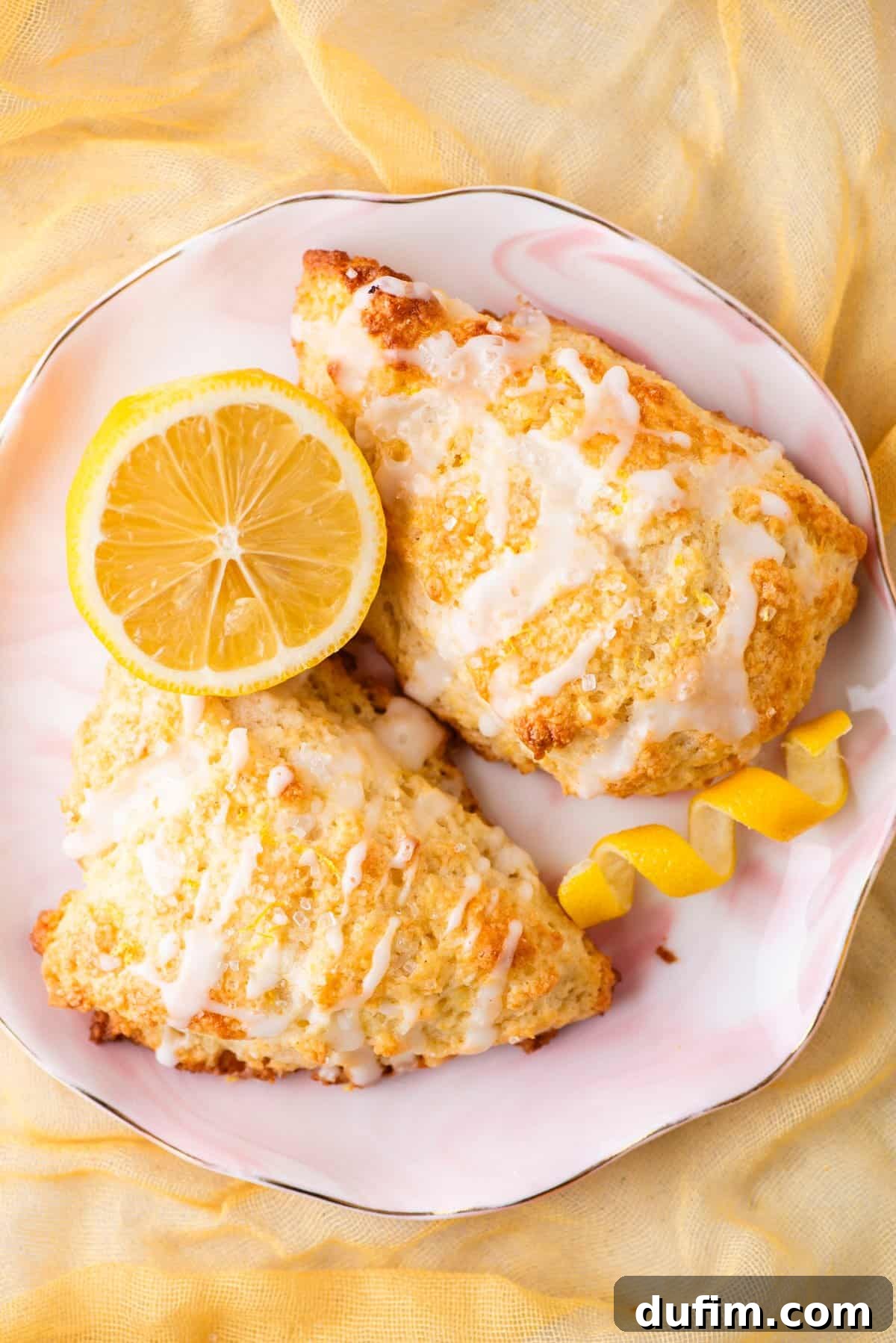 Two elegantly glazed gluten-free lemon scones on a white plate with a pink rim, garnished with a fresh lemon slice and a lemon twist. The scene is set against a soft yellow fabric, highlighting their inviting appearance.