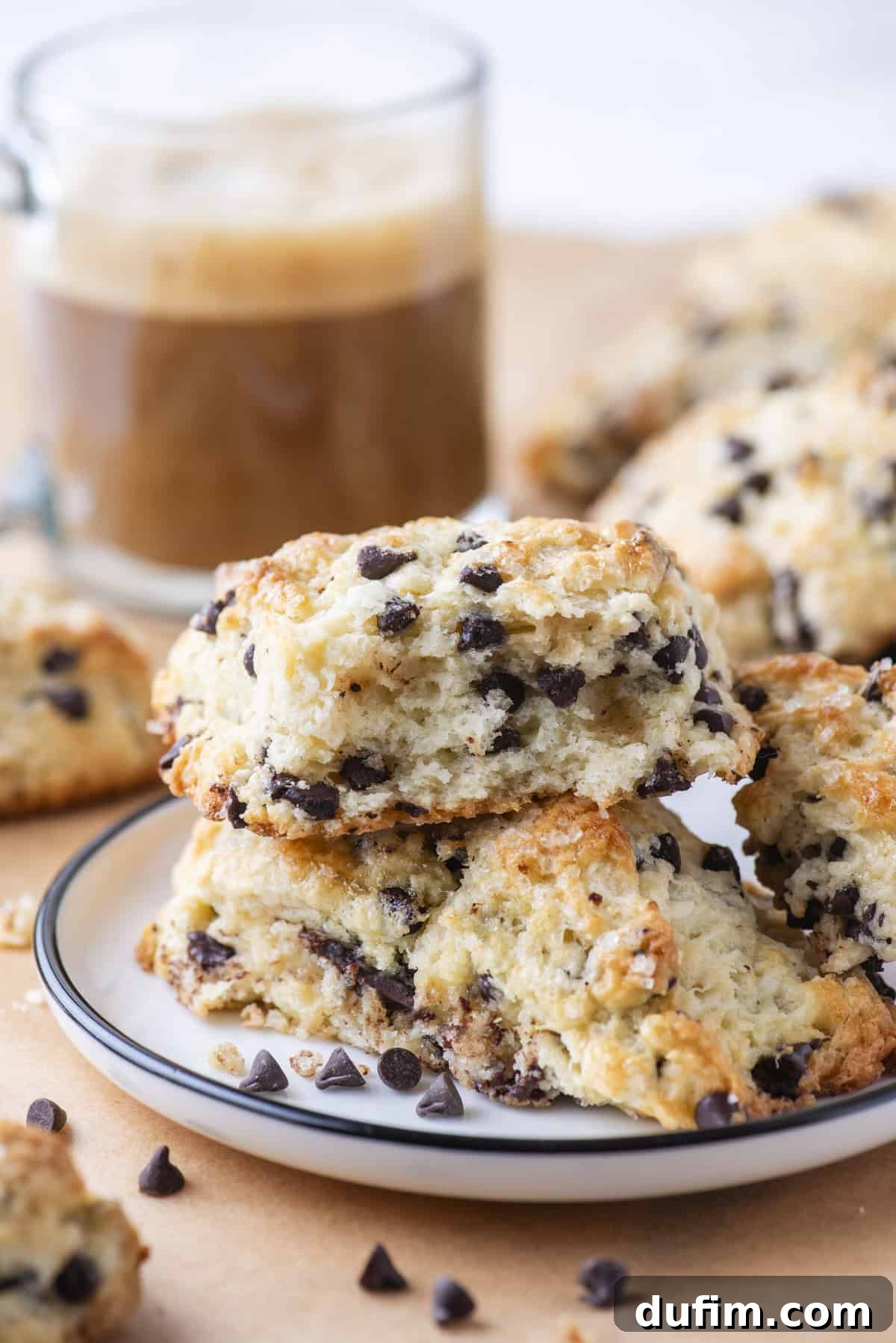 A stack of chocolate chip gluten-free scones on a white plate with a black rim, with more scones and scattered chocolate chips in the background. A blurred coffee cup adds to the cozy atmosphere.