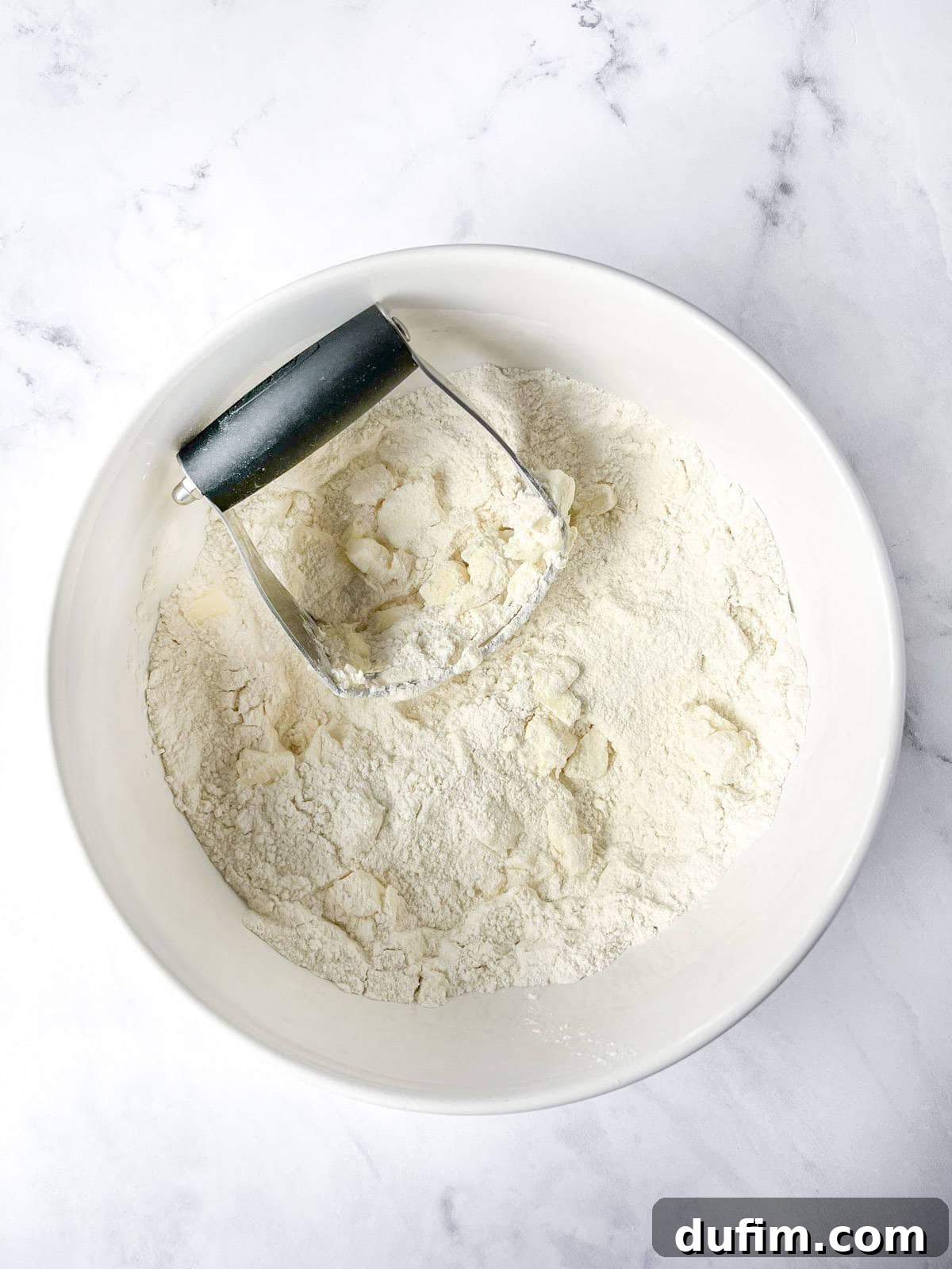 A white bowl containing gluten-free flour and small chunks of cold butter sits on a marble surface, with a pastry cutter resting inside the bowl, indicating the next step in scone preparation.