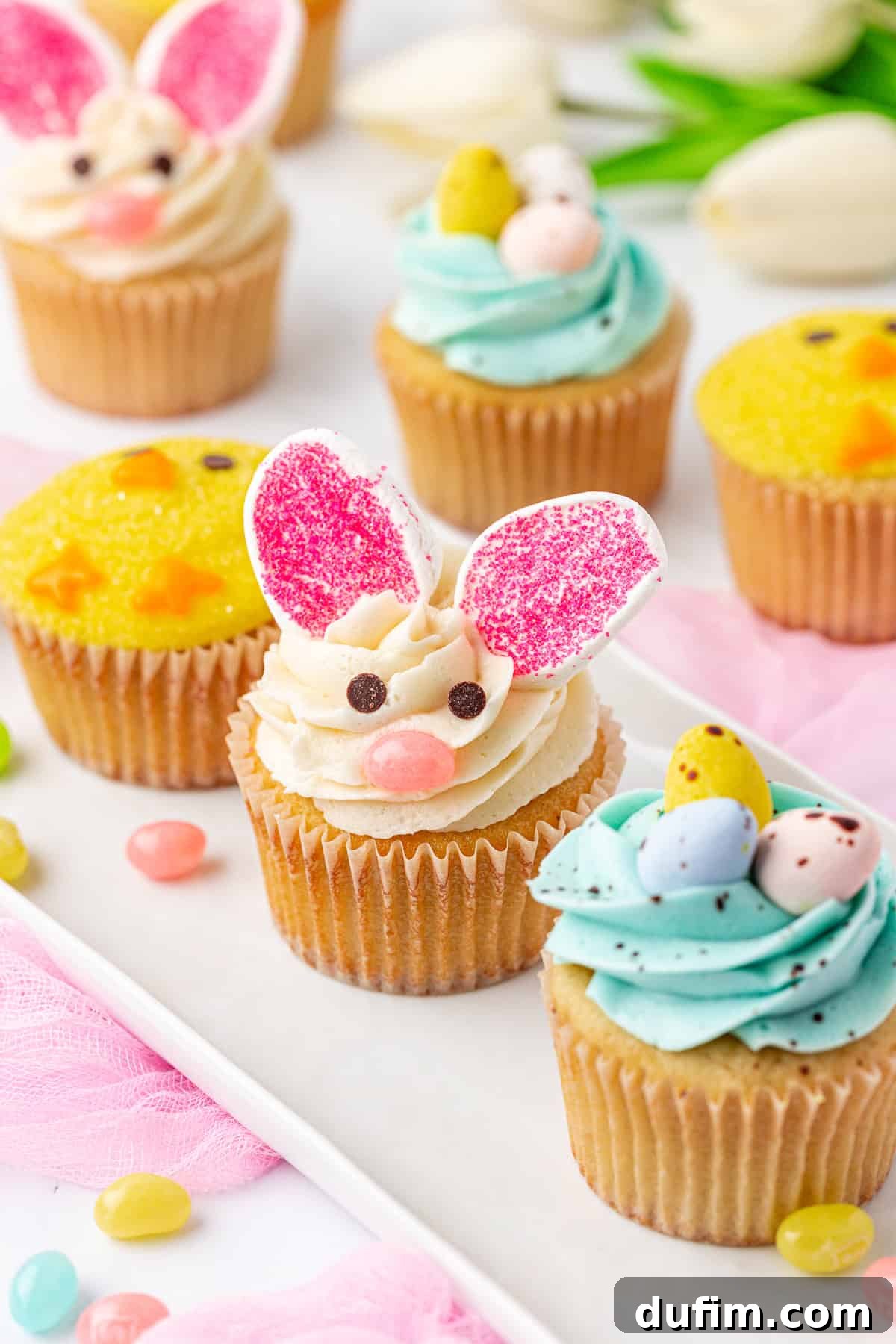 A platter of decorated cupcakes, including one with bunny ears and face made of frosting, and others topped with pastel-colored eggs and yellow sprinkles. Jellybeans and flowers are scattered around for a festive spring theme.