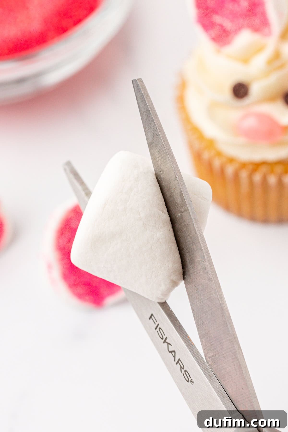 A pair of scissors cutting a white marshmallow, with a cupcake decorated like a bunny and a plate of pink treats in the background.