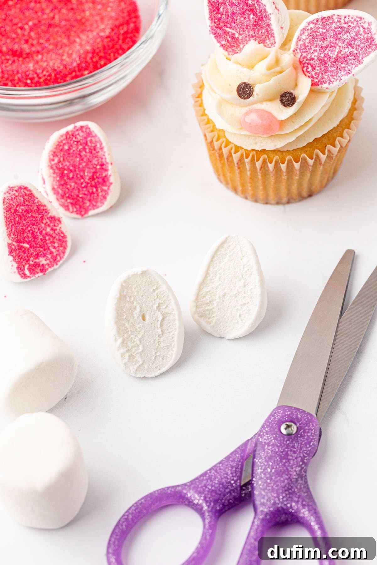 A cupcake decorated as a bunny with pink ears and a face sits near marshmallow halves, pink sugar, a bowl of sprinkles, and purple scissors on a white surface.