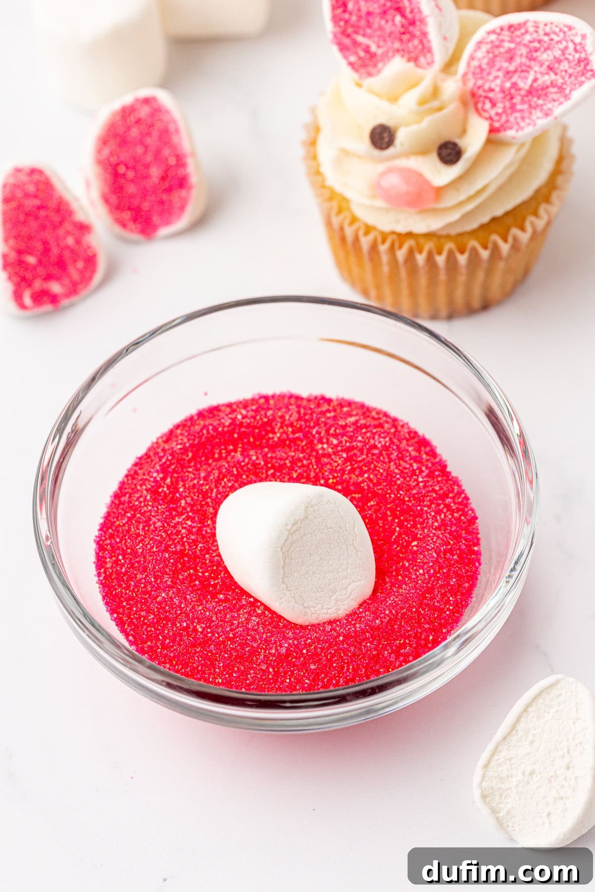 A bowl of pink sugar crystals with a marshmallow being dipped in it, surrounded by marshmallow pieces, and a cupcake decorated like a bunny with frosting, candy decorations, and marshmallow ears.