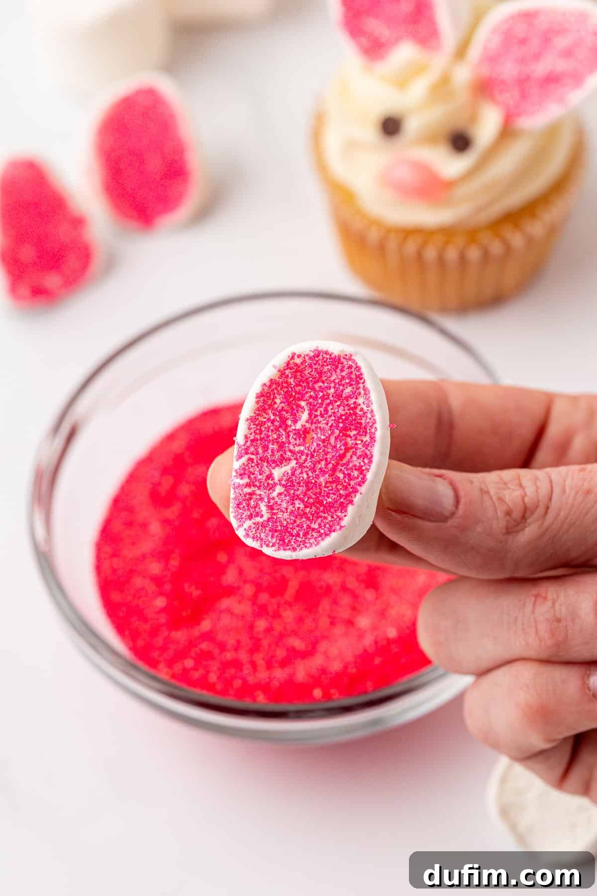 A hand holds a marshmallow half dipped in pink sugar over a bowl of pink sugar, with a bunny-themed cupcake and more pink-dipped marshmallow pieces in the background.