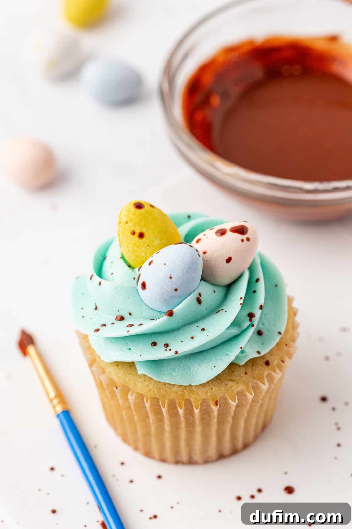 A cupcake with blue frosting is topped with three pastel candy eggs and chocolate speckles. A paintbrush and a bowl of melted chocolate sit nearby on a white surface.