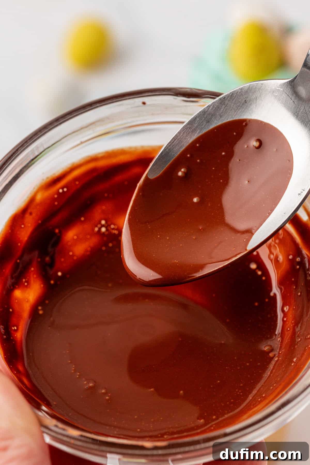 A close-up of a spoon lifting smooth, melted chocolate from a small glass bowl filled with glossy chocolate sauce. The background is softly blurred.